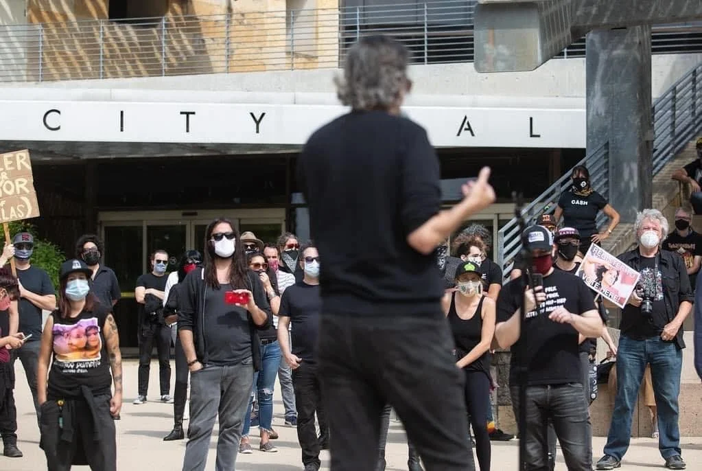 A group of protesters, mostly wearing masks, gather outside a building labeled 'City Hall.' One person is speaking into a microphone, while others hold signs and listen.