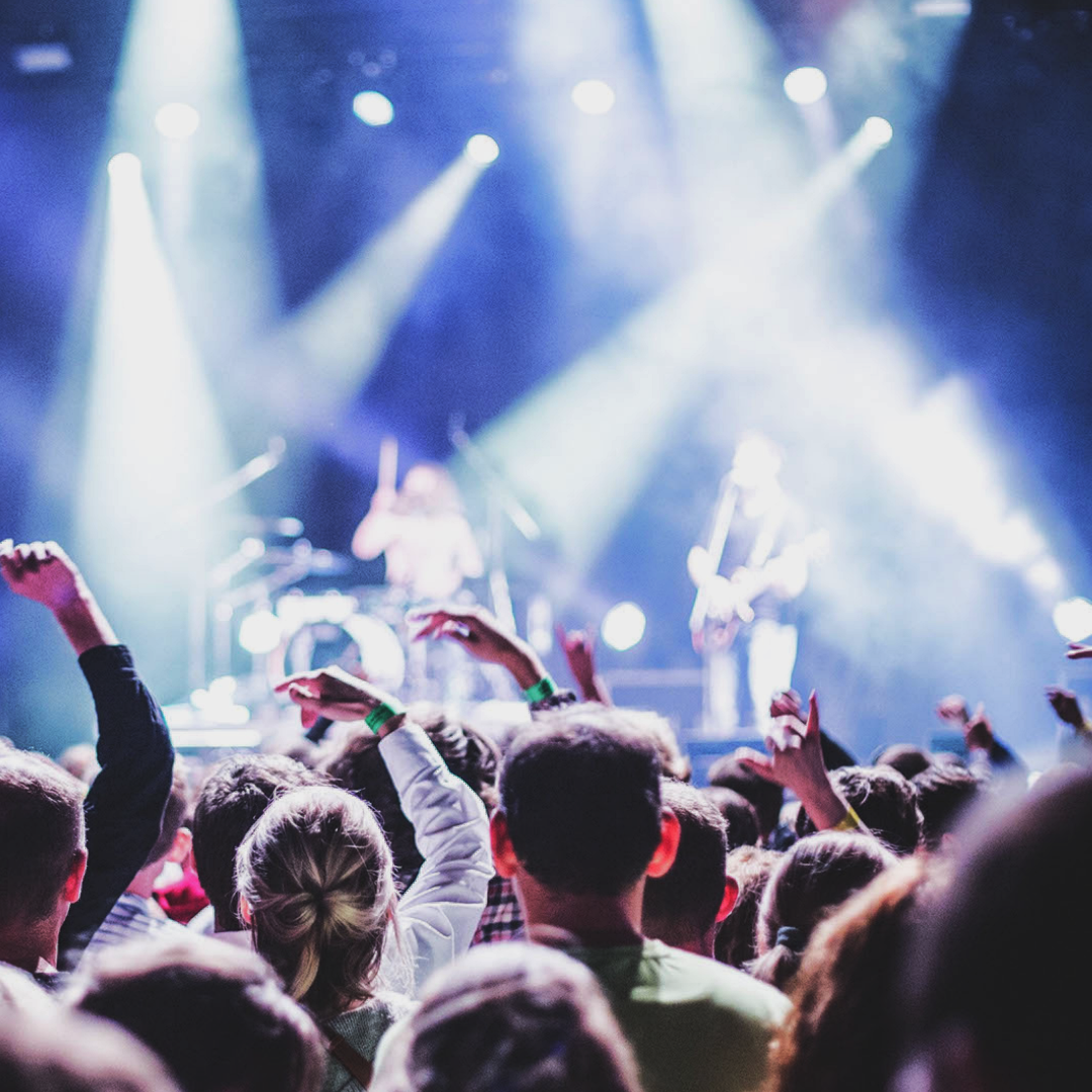Crowd at a concert with hands raised, stage with bright lights, and performers playing instruments in the background.