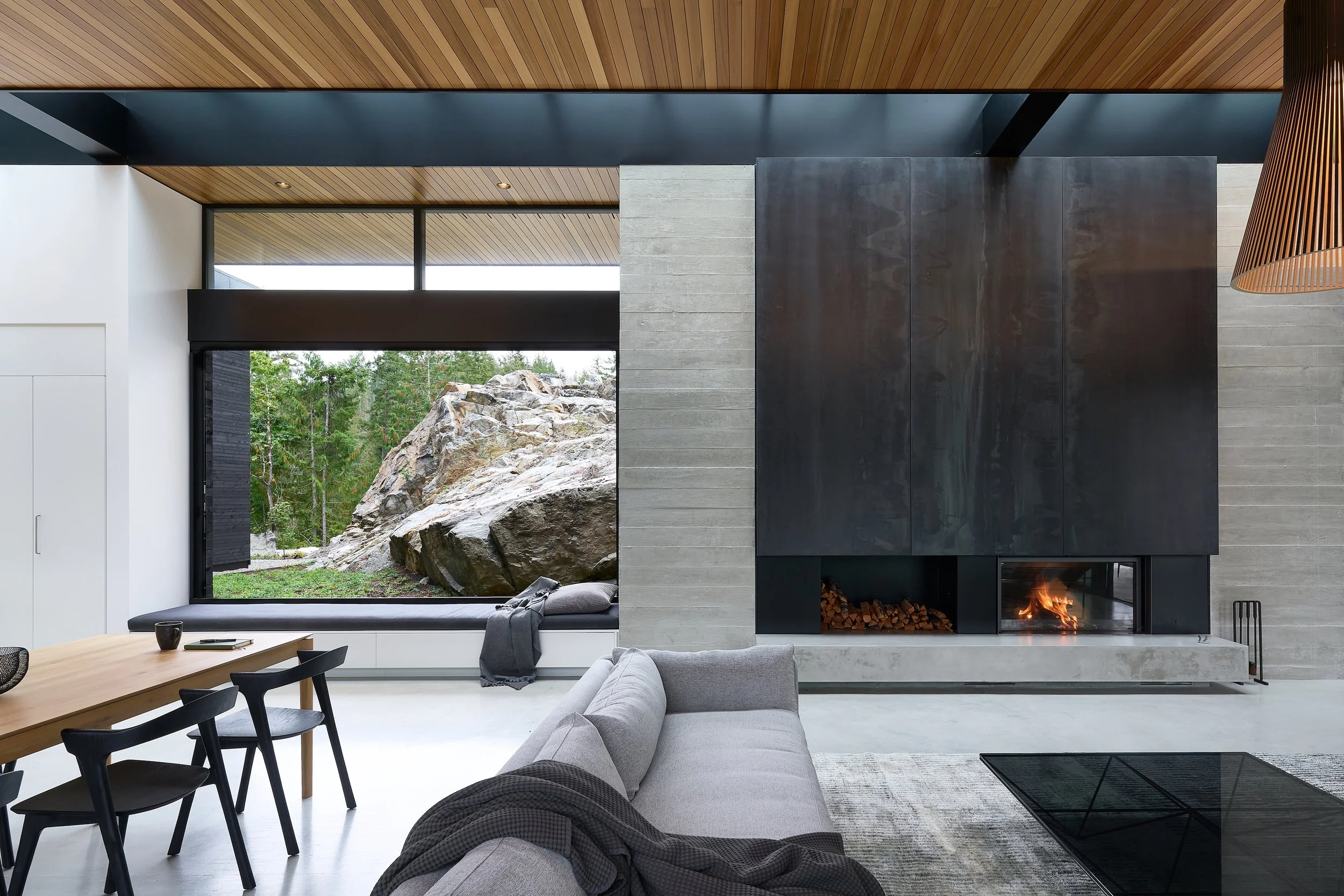 Modern living room with large window view of outdoor rocks and trees, gray sofa, black chairs, wooden dining table, fireplace, and ceiling with wood paneling.