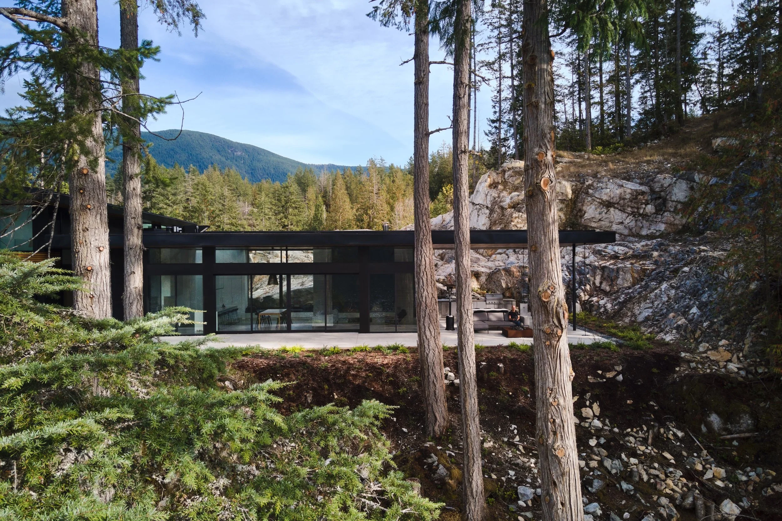 A modern glass house nestled among tall trees in a forested area, with mountain in the background and a patio with outdoor furniture in the foreground.