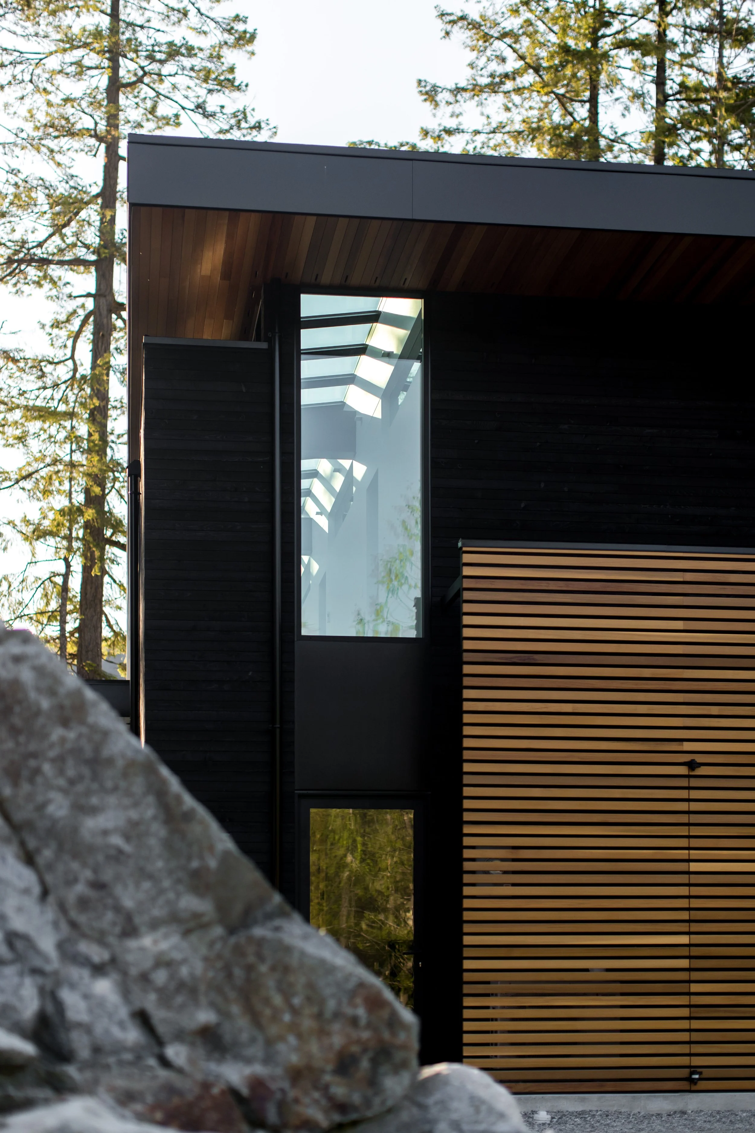 Close-up view of a modern house with black and wood exterior, large vertical window reflecting trees, surrounded by rocks and trees in the background.
