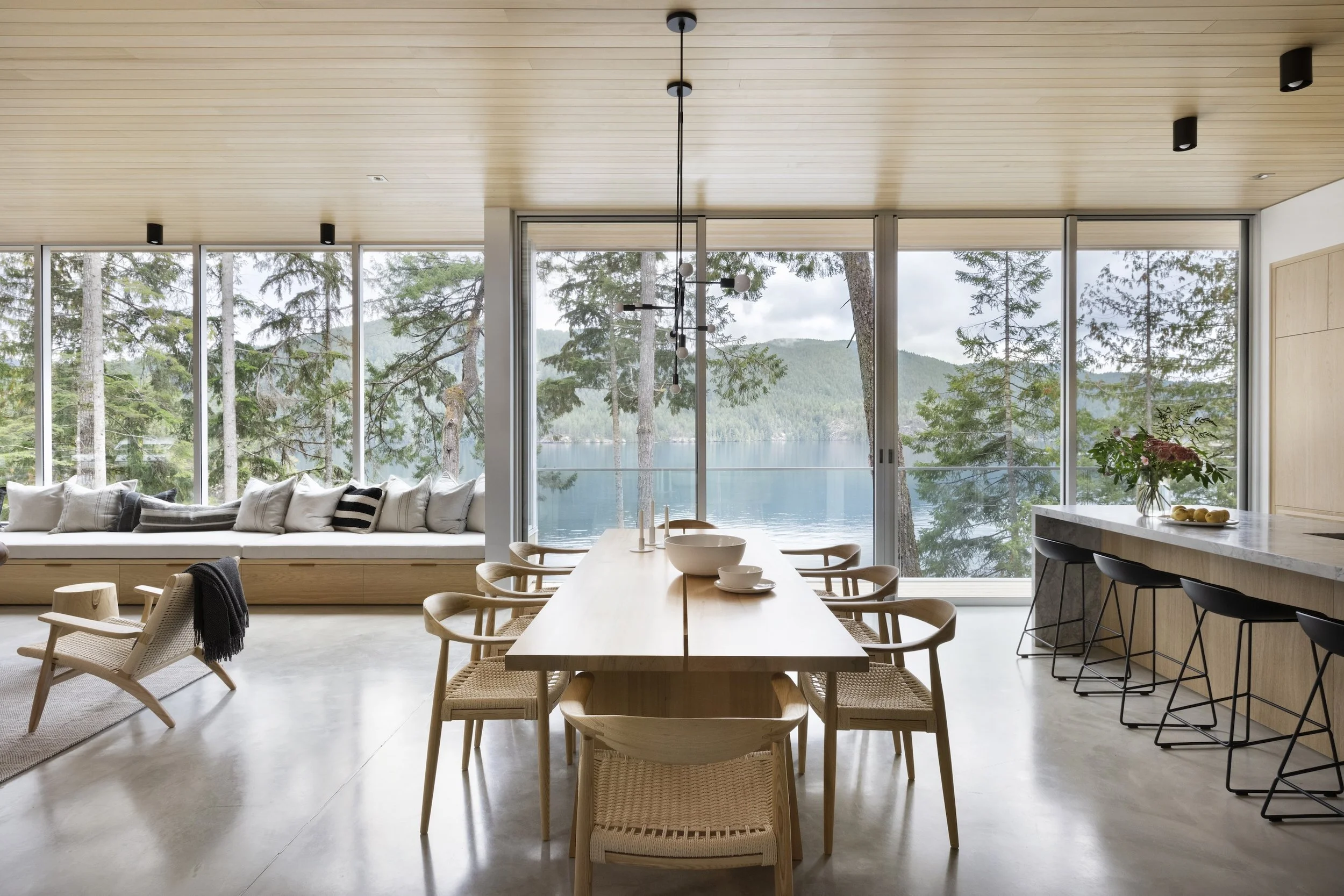 Modern dining and living room with large windows overlooking a lake and forest, wooden ceiling, minimalist furniture, and neutral color palette.
