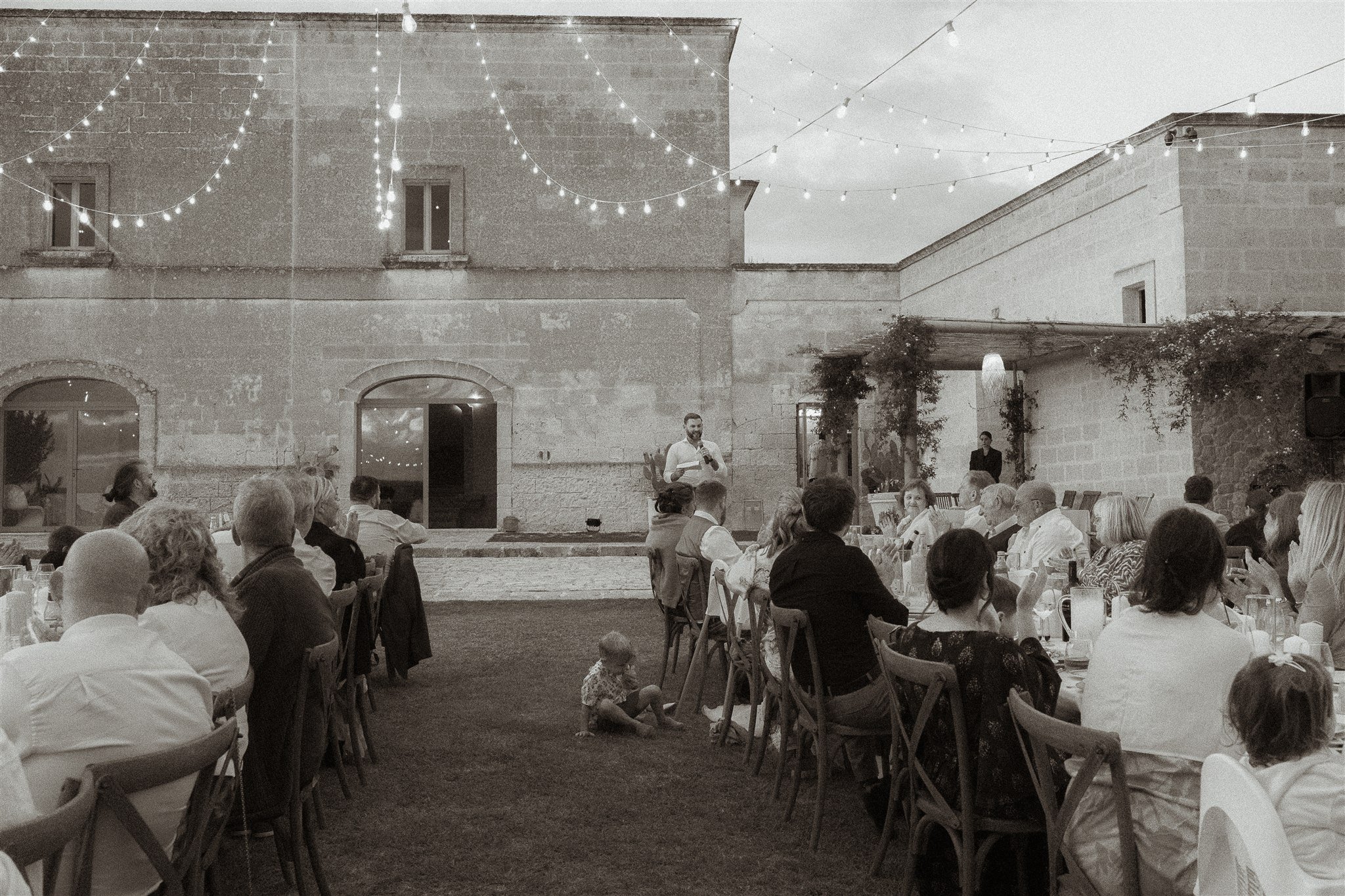 Outdoor gathering at night with people seated at tables, a man speaking at a microphone, string lights overhead, stone building in background, a small child sitting on grass in foreground