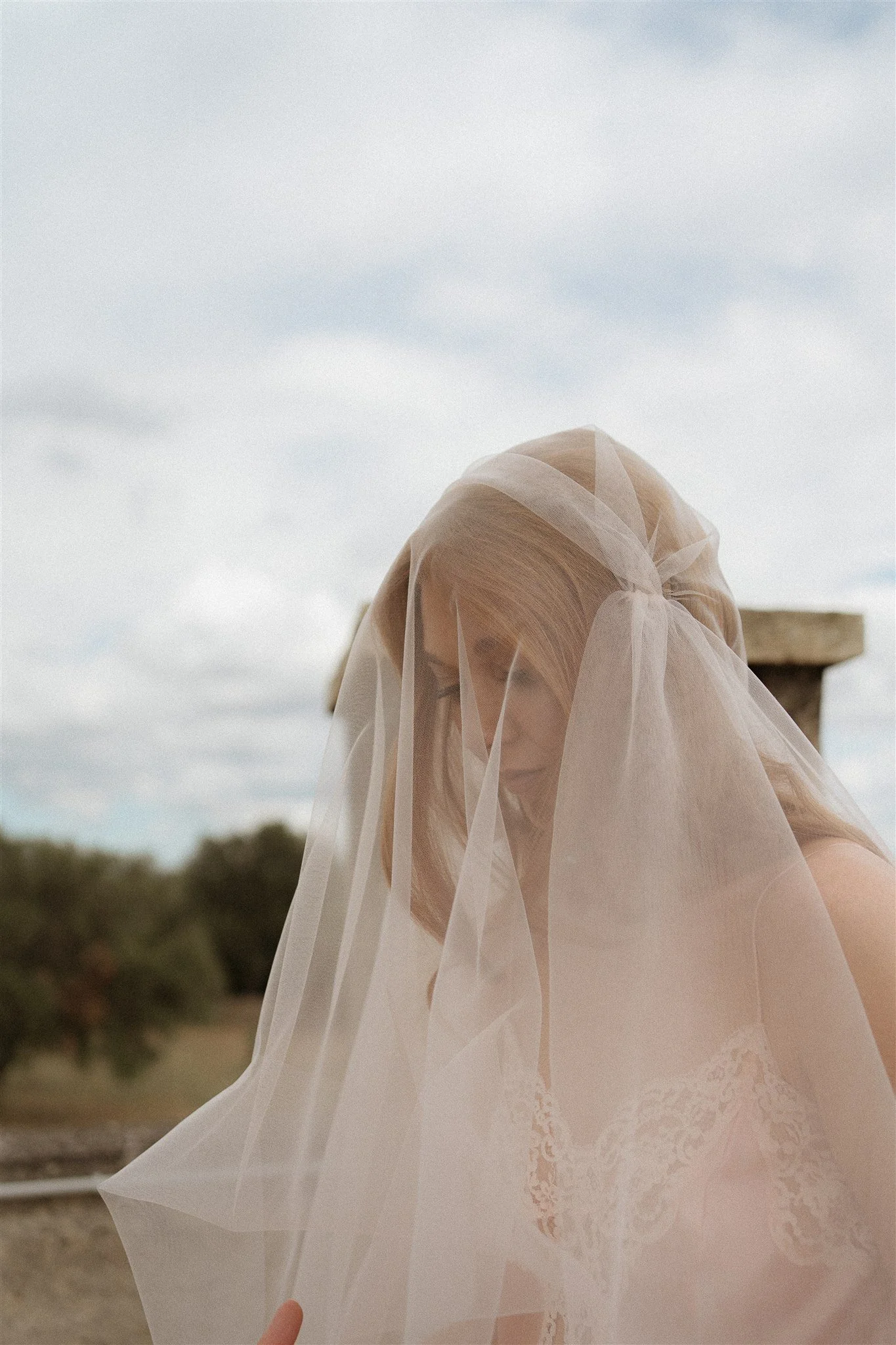 A woman with long blonde hair, dressed in a lace outfit, has a sheer veil over her face and is smiling outdoors, with trees and a cloudy sky in the background.