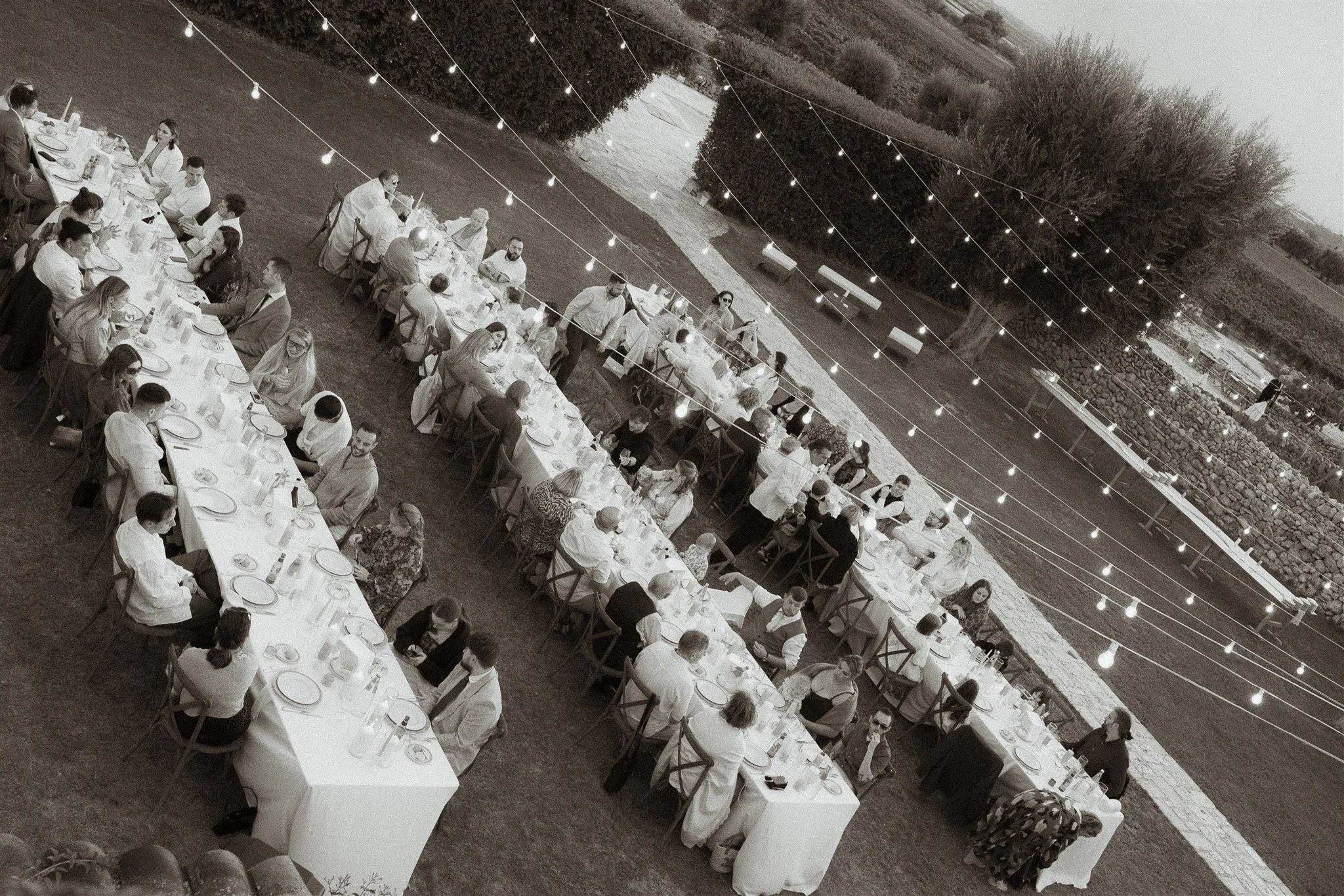 Outdoor dinner set up with multiple long tables surrounded by people, string lights overhead, and a grassy yard with trees and a stone wall in the background.