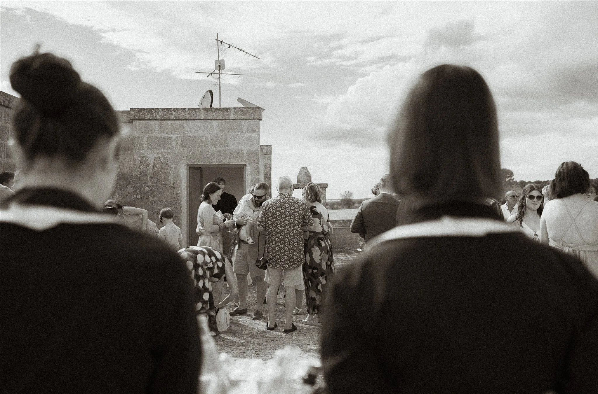 People gathered on a terrace outdoors, with two women in the foreground facing away, watching a group of people in the middle. The scene appears to be a casual social event, with a stone building and TV antennas visible under a partly cloudy sky.