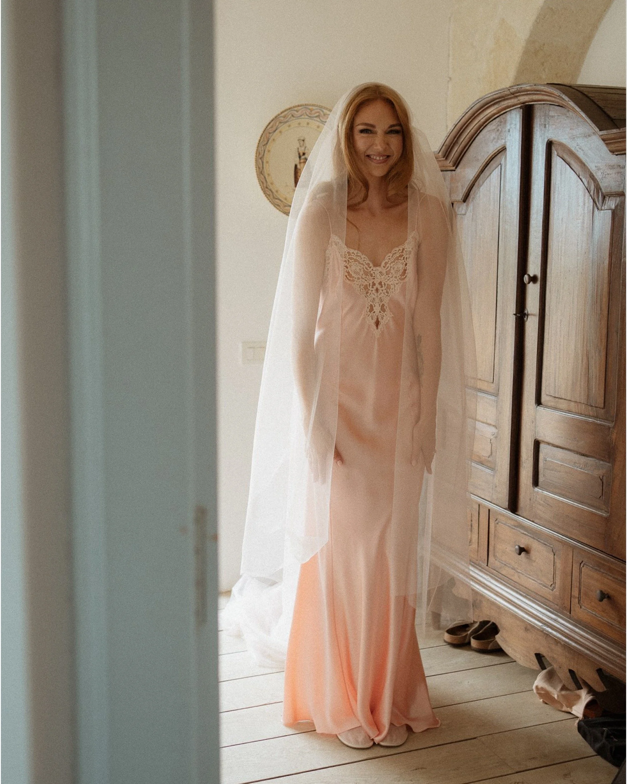 A woman in a pink lace-trimmed nightgown and a veil, smiling and standing in a room with wooden furniture.
