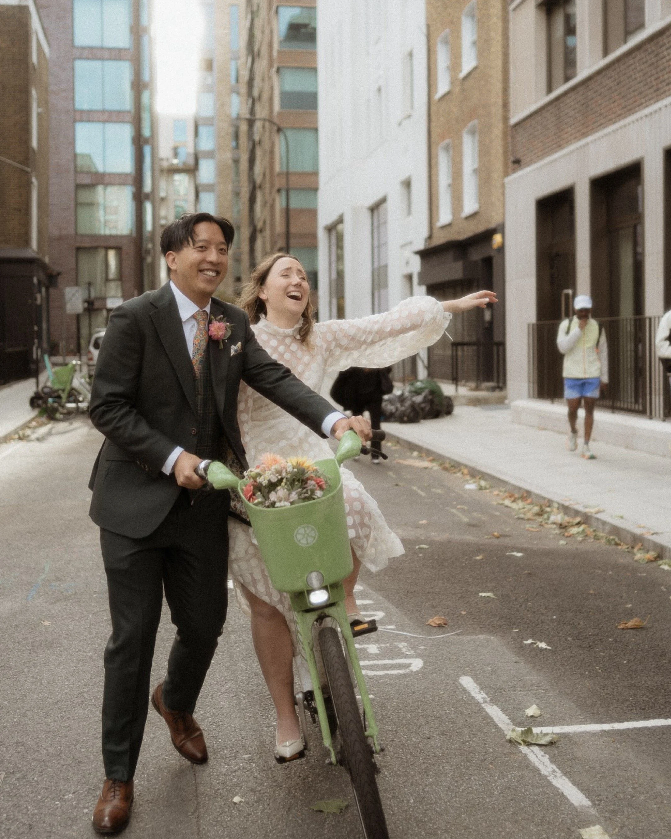 A man and woman in formal attire enjoying a bike ride on a city street, both smiling and appearing very happy, with buildings in the background.