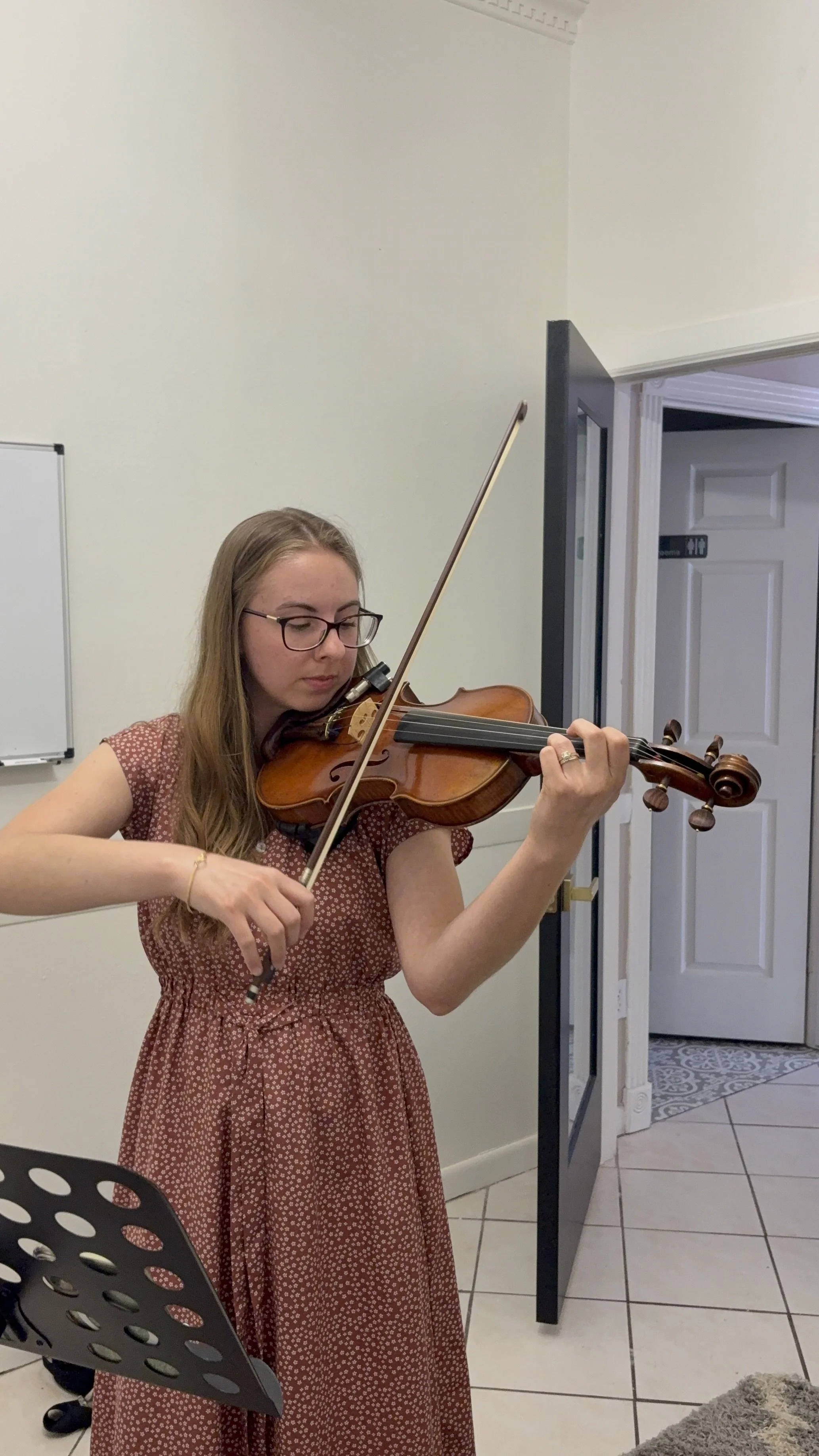 A woman with glasses playing a violin indoors, wearing a red dress with white polka dots, in front of a music stand and near a doorway.