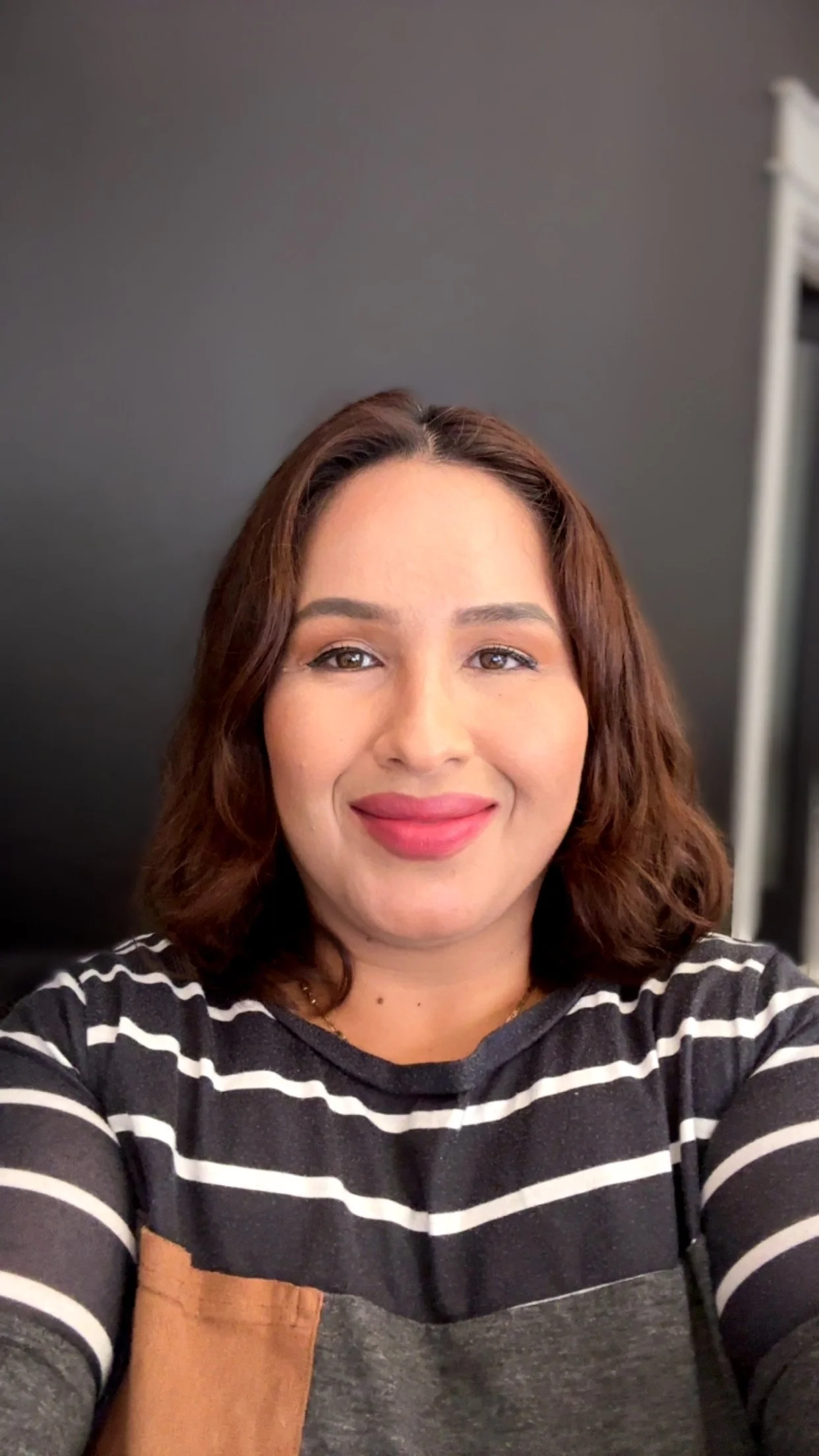 A woman with shoulder-length brown hair taking a selfie in front of a black background, wearing a black and white striped shirt, smiling softly.