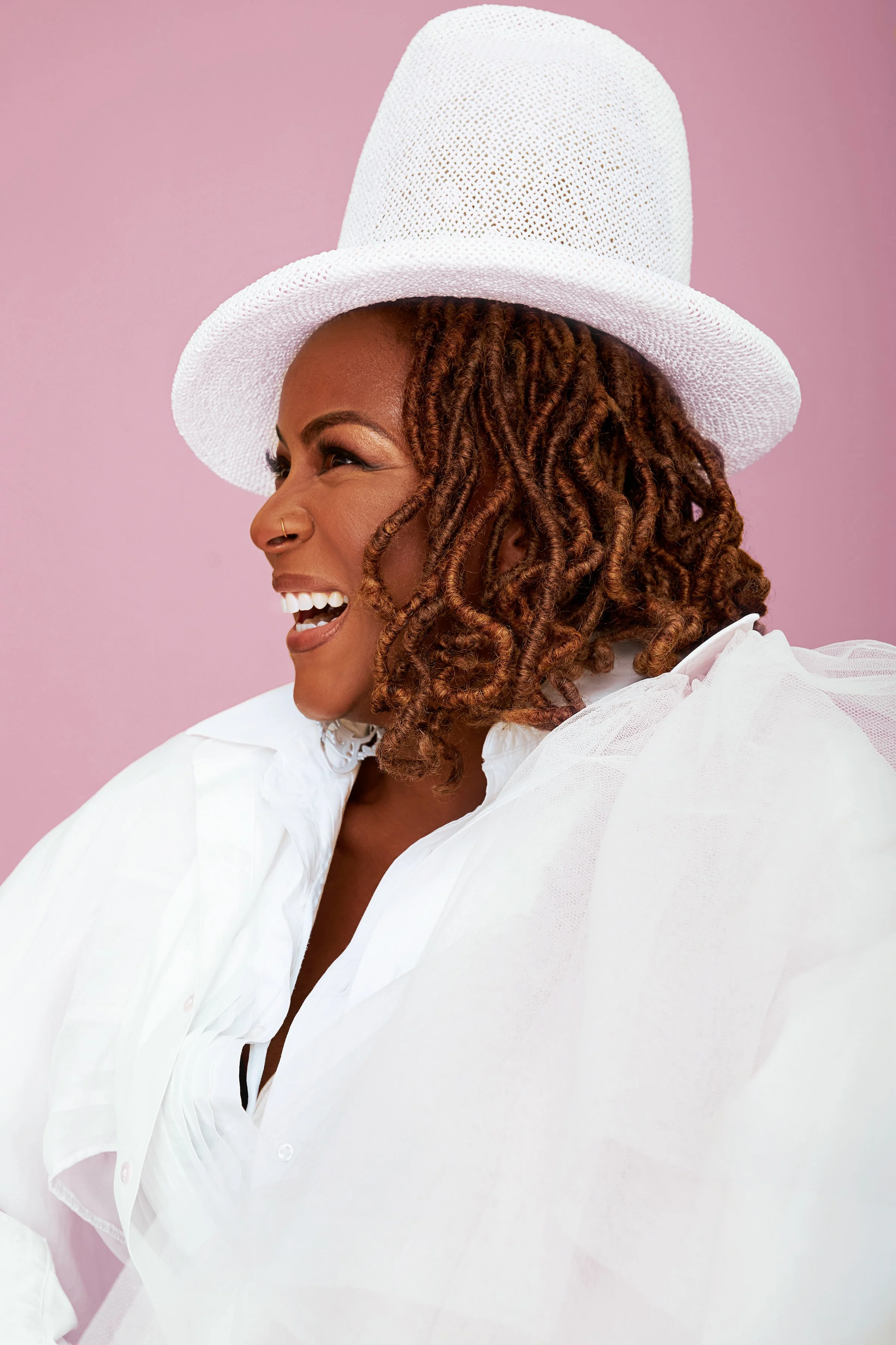 A black feme person wearig a tall, white, brimmer hat and a fluffy white top on a pink background. She is smiling wide and facing left.