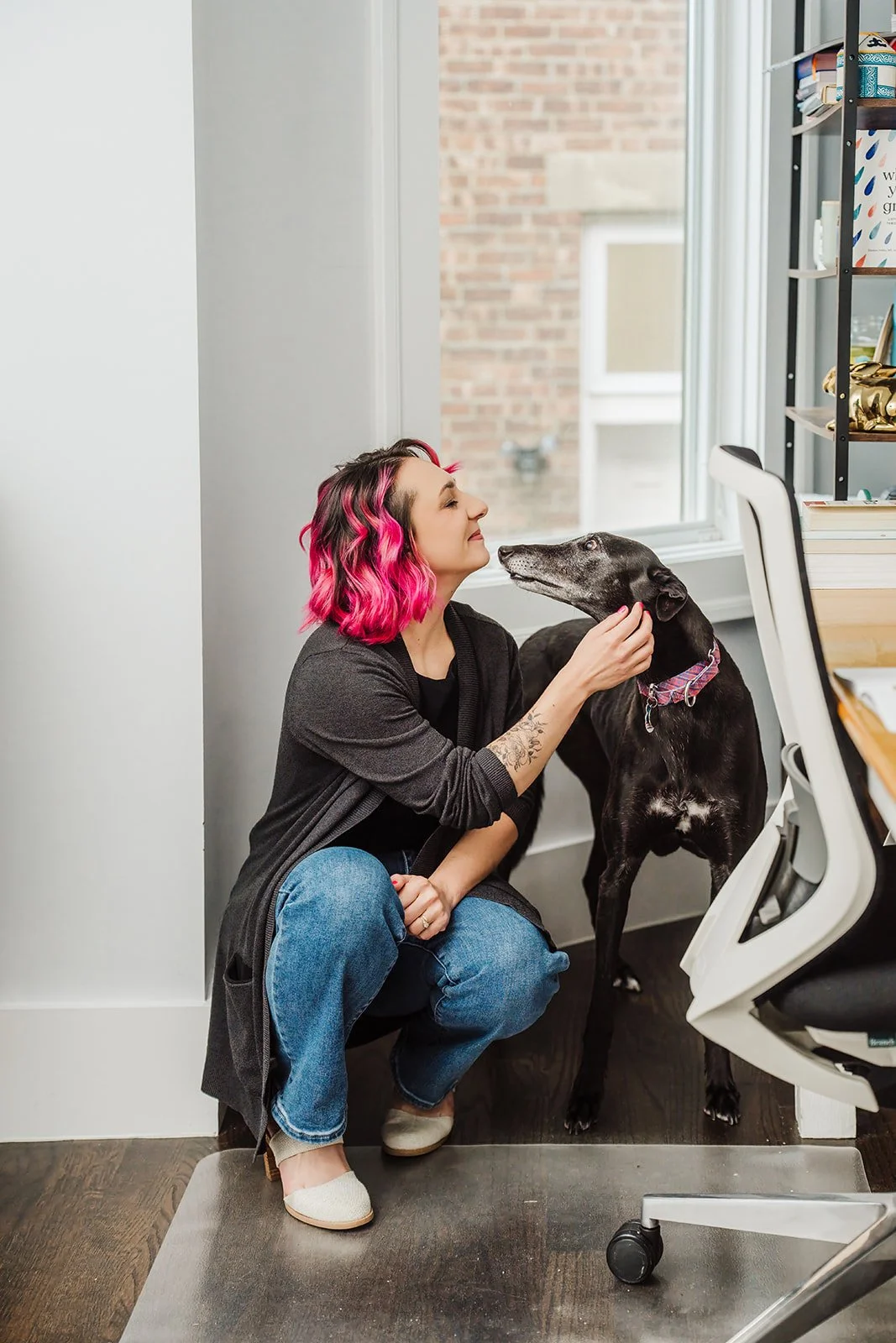 A photo of a white woman with pink hair, dressed in a dark gray sweater, black shirt, jeans, and white shoes, crouching down next to a black greyhound. They are both looking at each other.