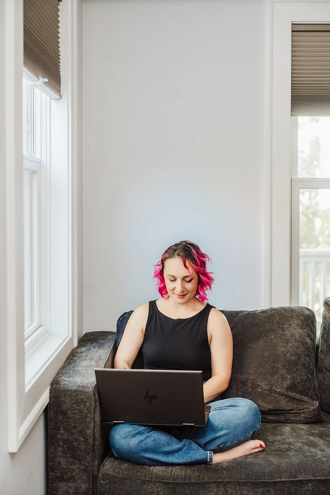 A white woman with pink hair, dressed in a black top and jeans, is sitting on a gray couch, looking down at the gray laptop in her lap.