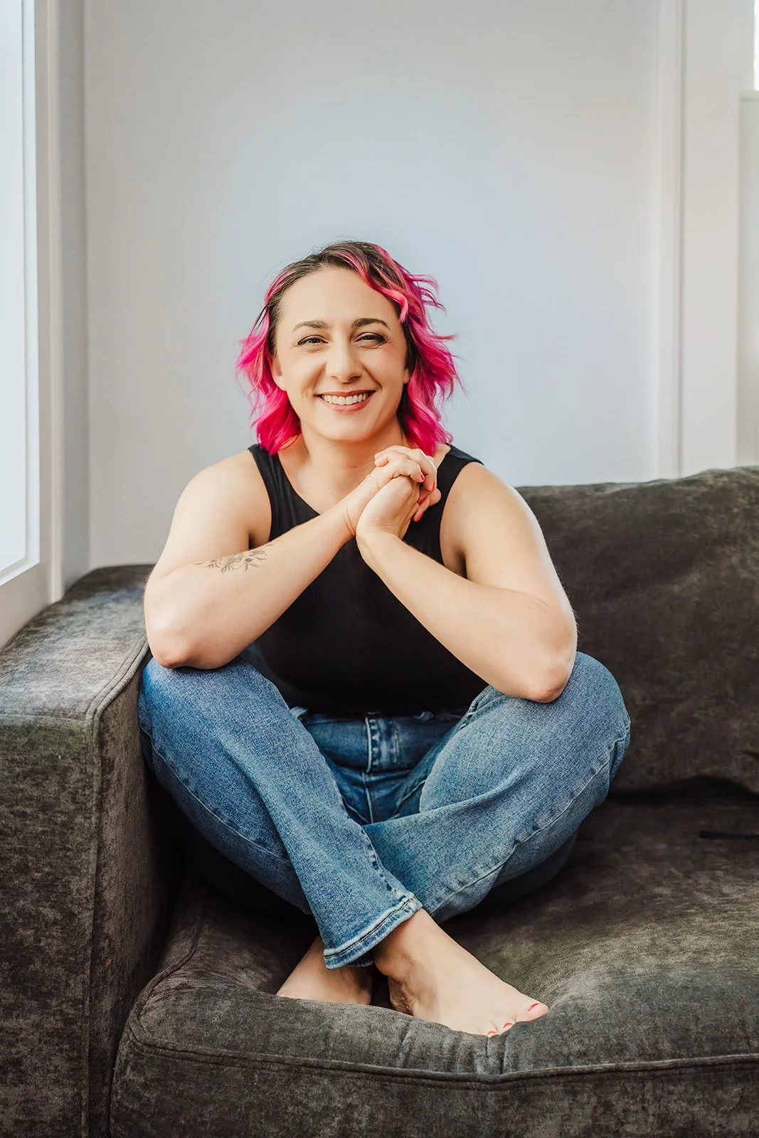 A photo of a white woman with pink hair, dressed in a black shirt and jeans. She is sitting on a gray couch, her hands clasped, elbows on her knees, smiling at the camera.