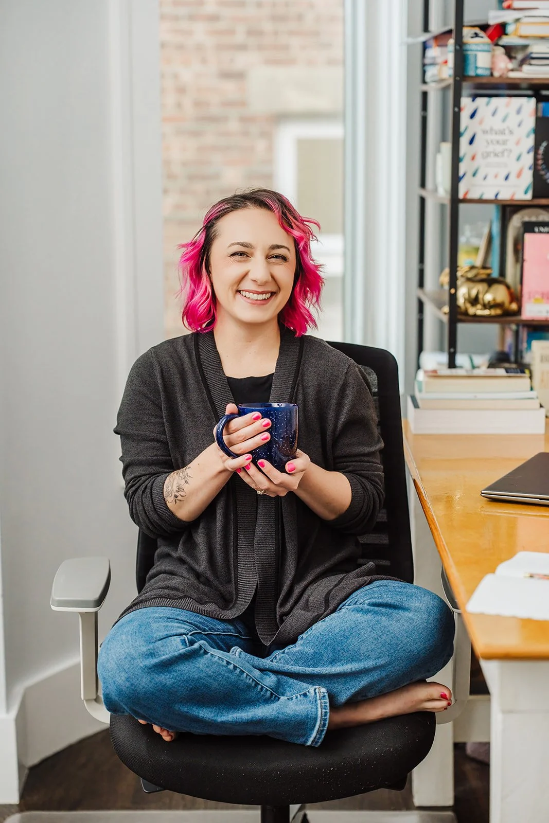 A photo of a white woman with pink hair, dressed in a dark gray sweater, black shirt, and jeans, sitting in an office chair at her desk with a window and bookcase behind her. She is smiling at the camera, holding a blue coffee mug in her hands.