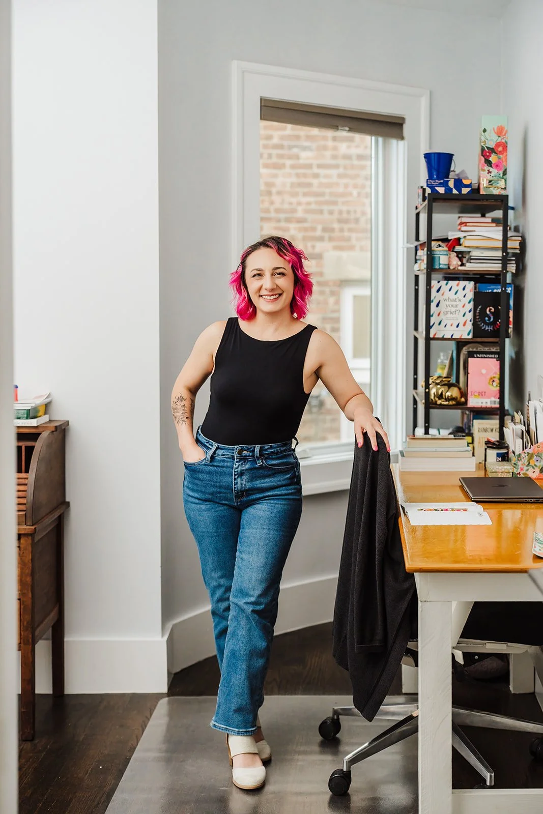 A photo of a white woman with pink hair, dressed in a black shirt, jeans, and white heeled shoes, standing in an office by her desk with a window and bookcase behind her. She is smiling at the camera.