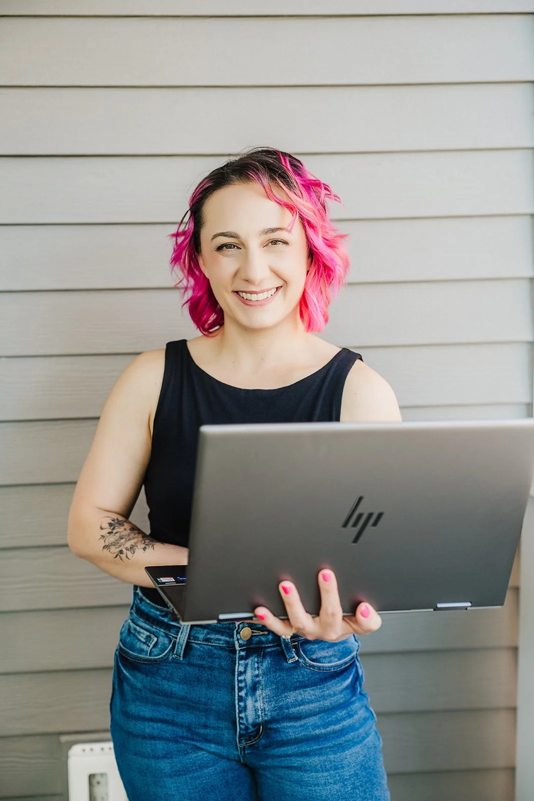 A photo of a white woman with pink hair wearing a black top and blue jeans, smiling at the camera against a gray backdrop with a laptop in her hands.