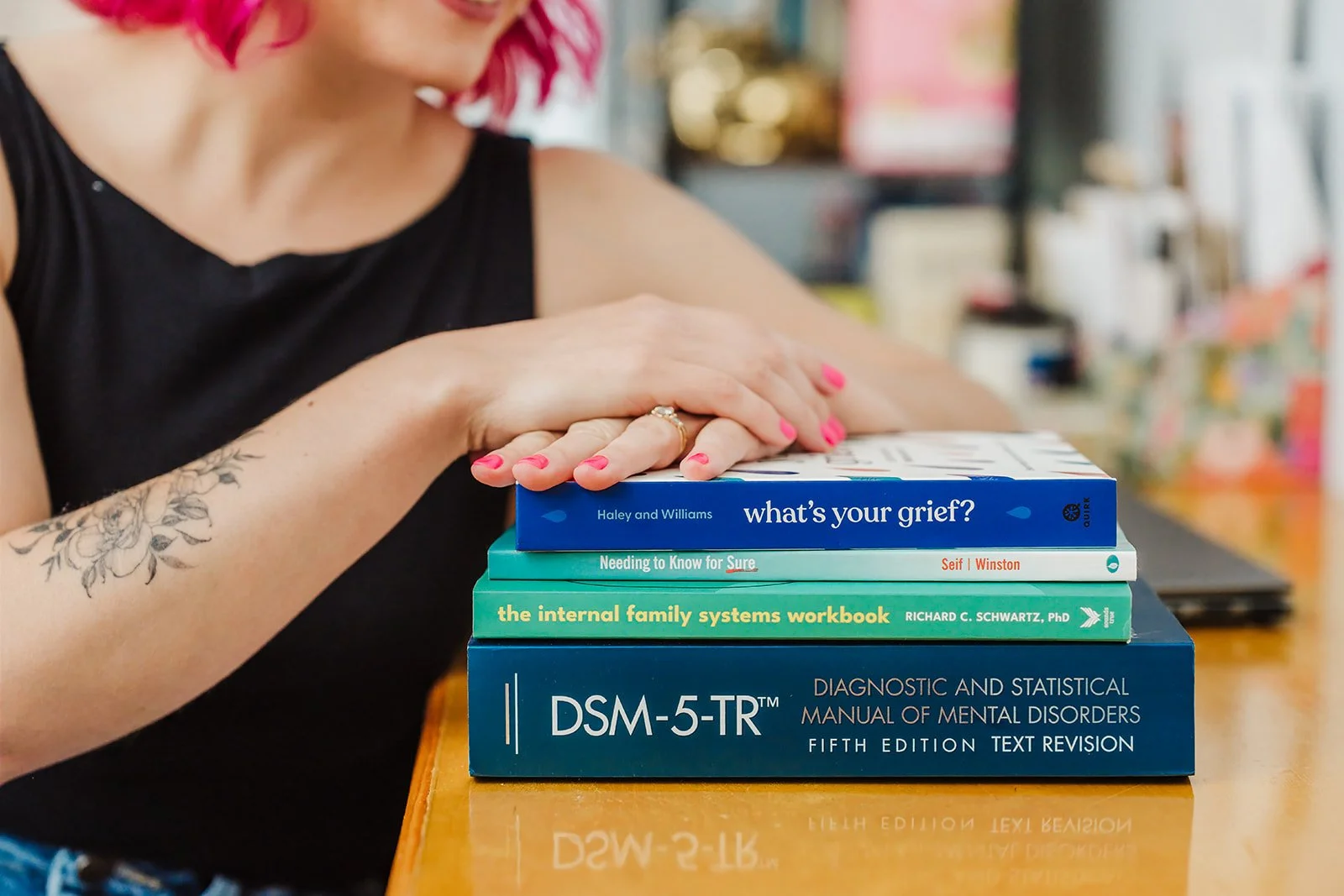A photo of a white woman's arms resting on a pike of books with pink nails visible, as well as a floral tattoo on her forearm.