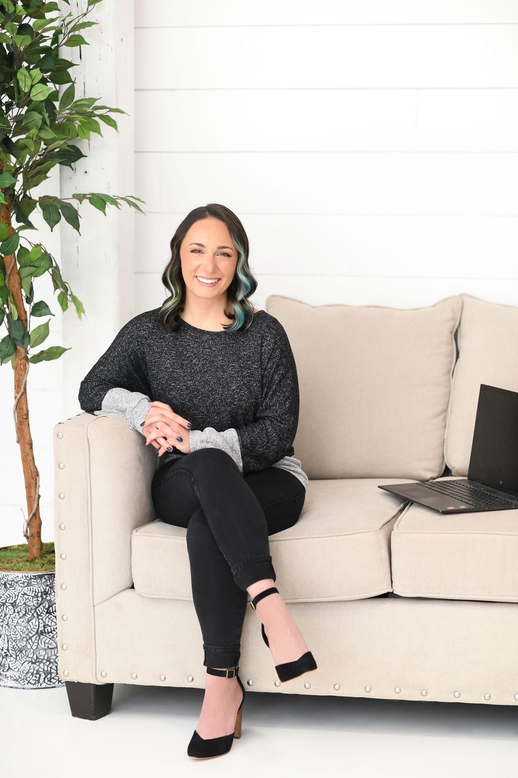 A picture of Sarah Kalny sitting on a cream colored couch with a plant to her right and laptop to her left. She is wearing a black and white shirt with black pants and black heeled shoes. Her dark hair has blue highlights.