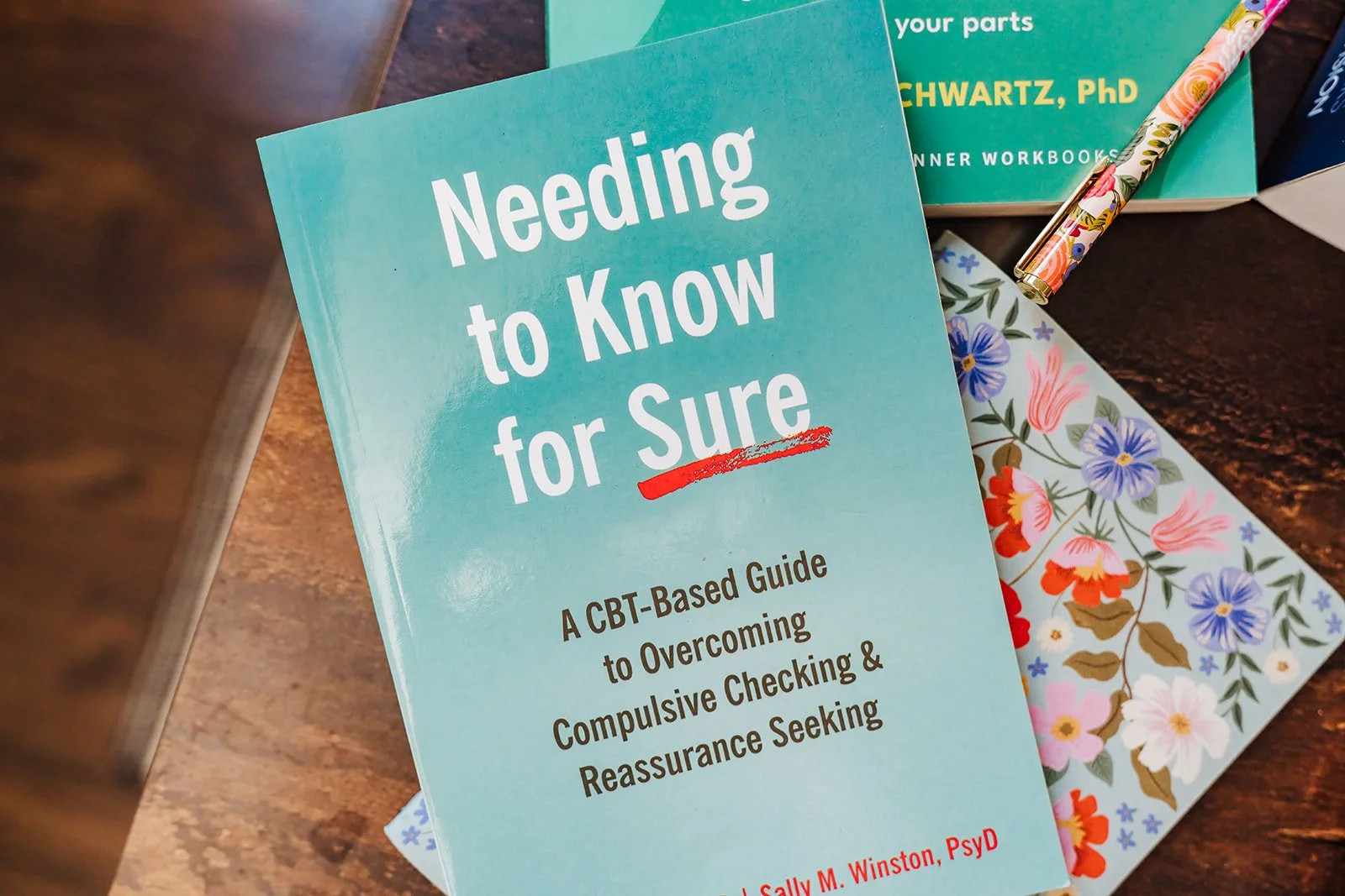 A picture of the book Needing to Know for Sure A CBT-Based Guide to Overcoming Compulsive Checking and Reassurance Seeking, on top of a floral notebook, placed on a wooden table.