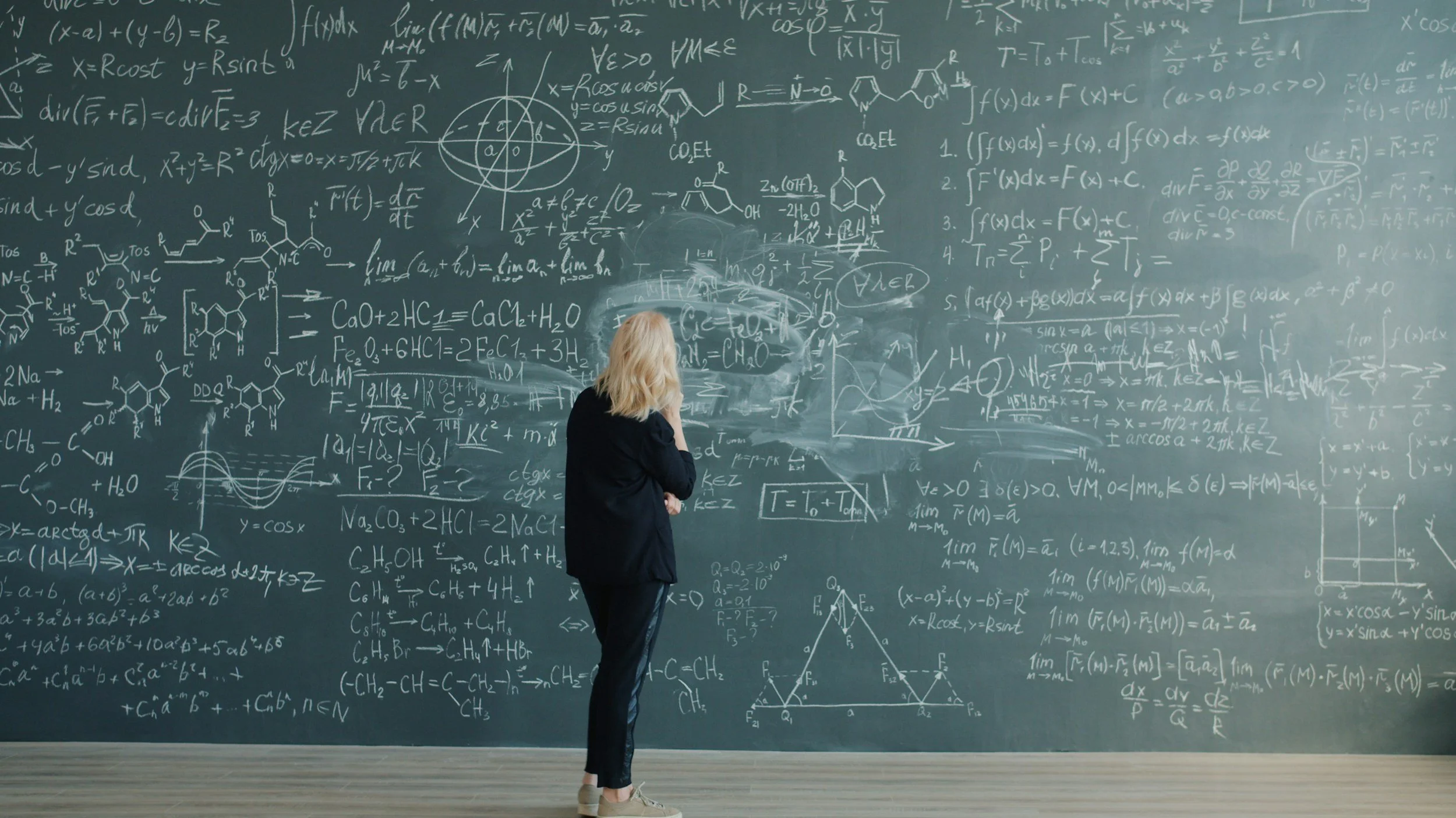 A woman with blonde hair, dressed in a dark blazer and pants, standing in front of a large chalkboard filled with complex mathematical and chemical equations, diagrams, and graphs.
