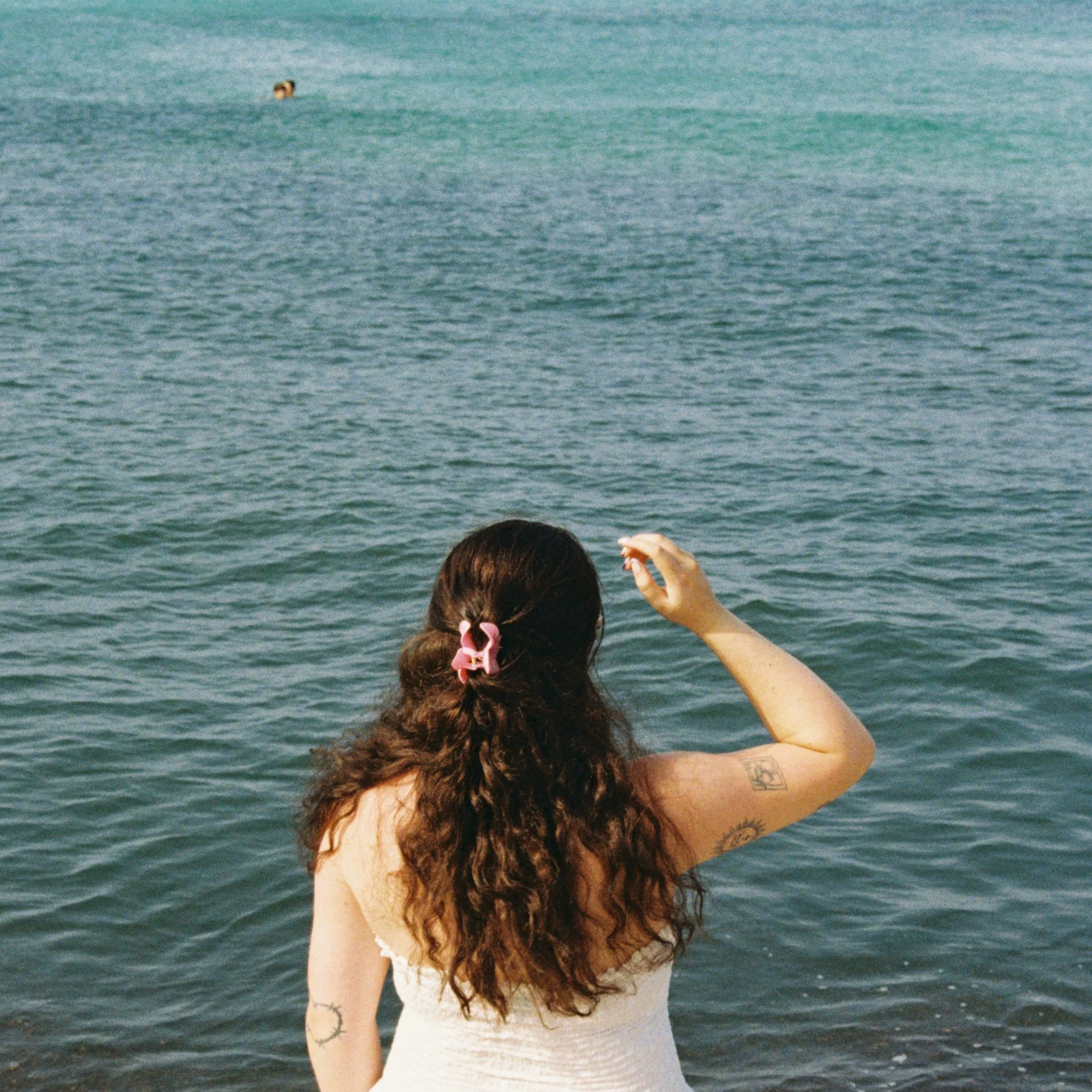Woman with long, curly hair with pink hair clip, standing by the water, partially visible from behind, wearing a white top, with tattoos on arm, facing the ocean.