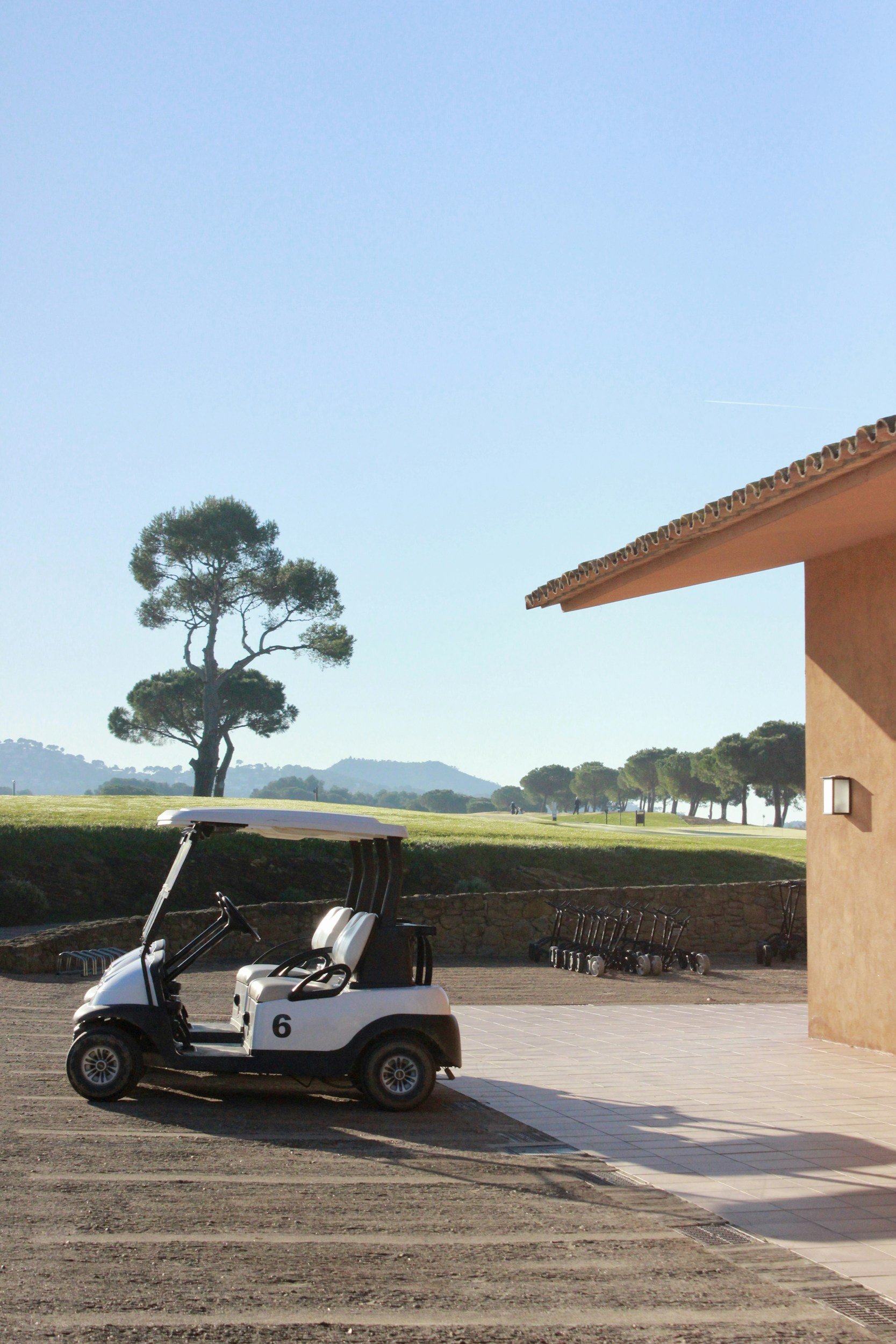 A golf cart parked beside a clubhouse at the edge of a fairway, with pines and low hills beyond.