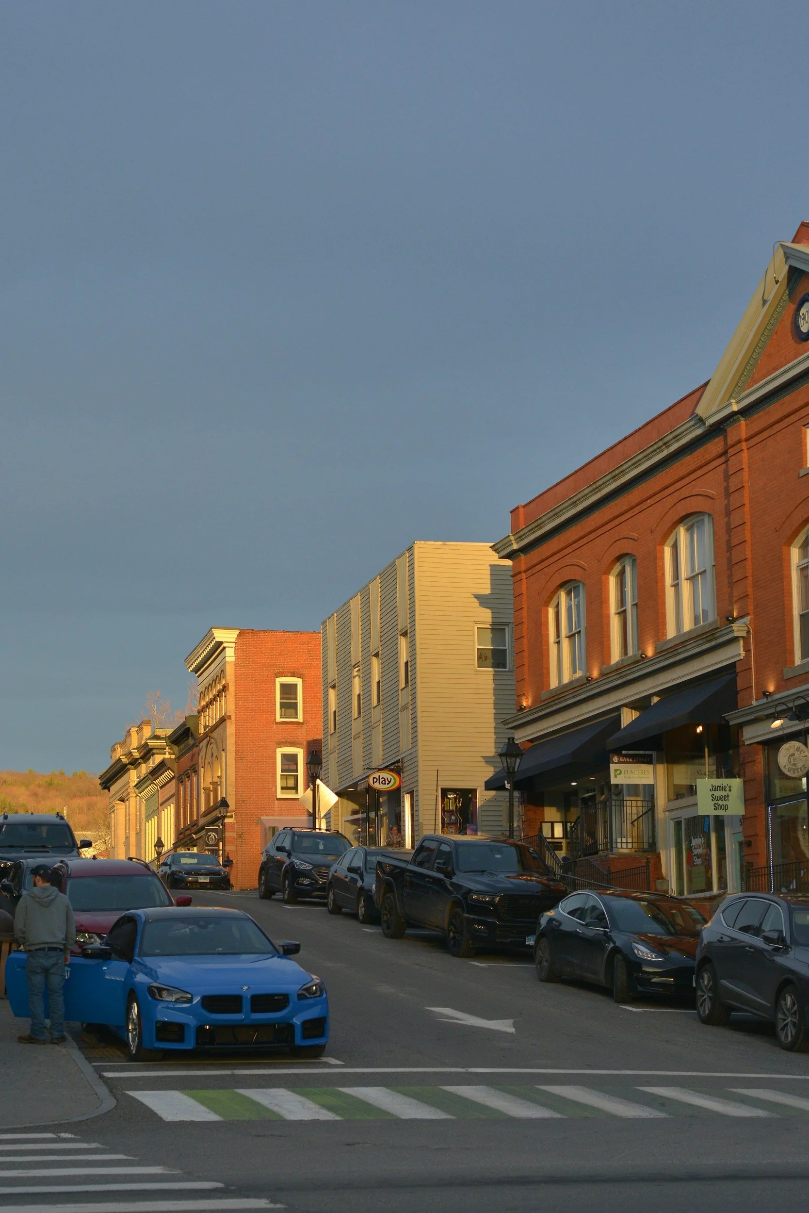 Brick storefronts and parked cars along a small-town main street in low evening sun.