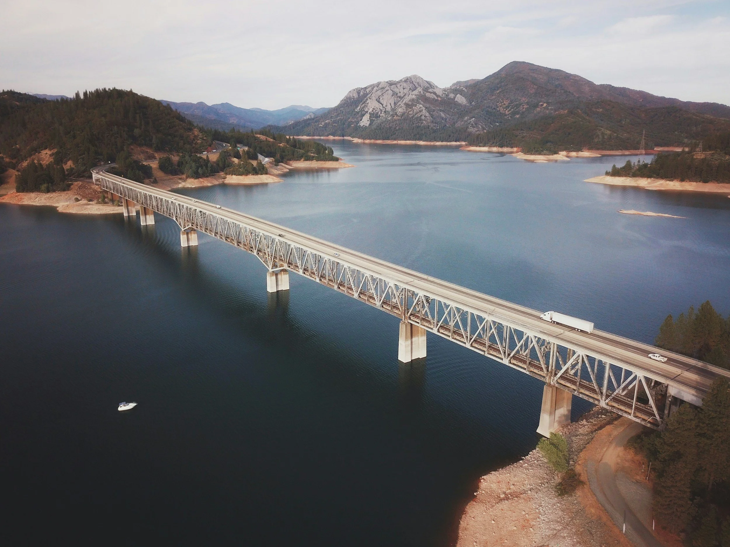 A long steel truss bridge carrying a highway across a wide blue lake, with pine-covered ridges behind and a small boat on the water.