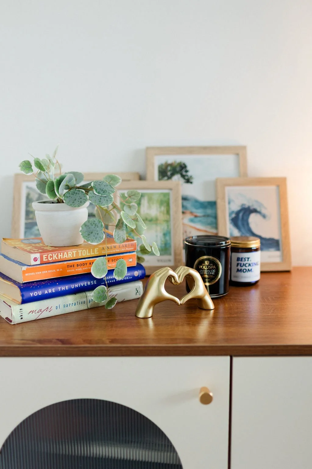 A wooden surface with a small potted plant, a stack of books, candles, framed pictures, and a gold sculpture forming a heart shape.