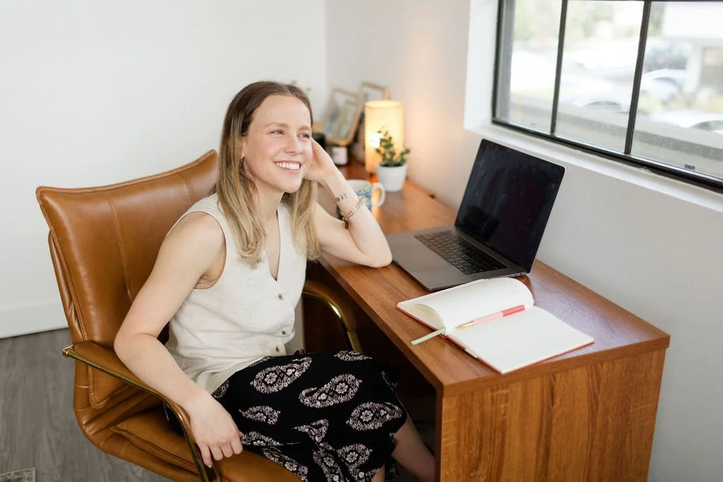 A woman sitting at a wooden desk near a window, smiling, with a laptop, an open notebook, and a pen on the desk, in a well-lit room.