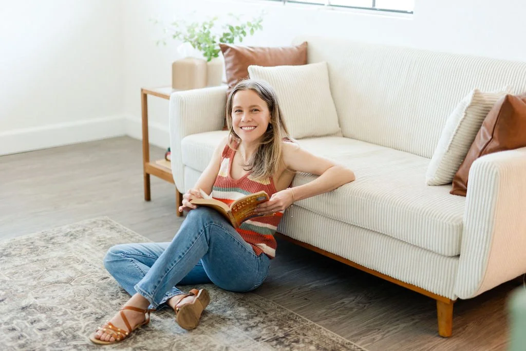 A smiling woman sitting on the floor with a book in her hand next to a white sofa with beige and brown pillows, in a bright living room with a plant on a side table.