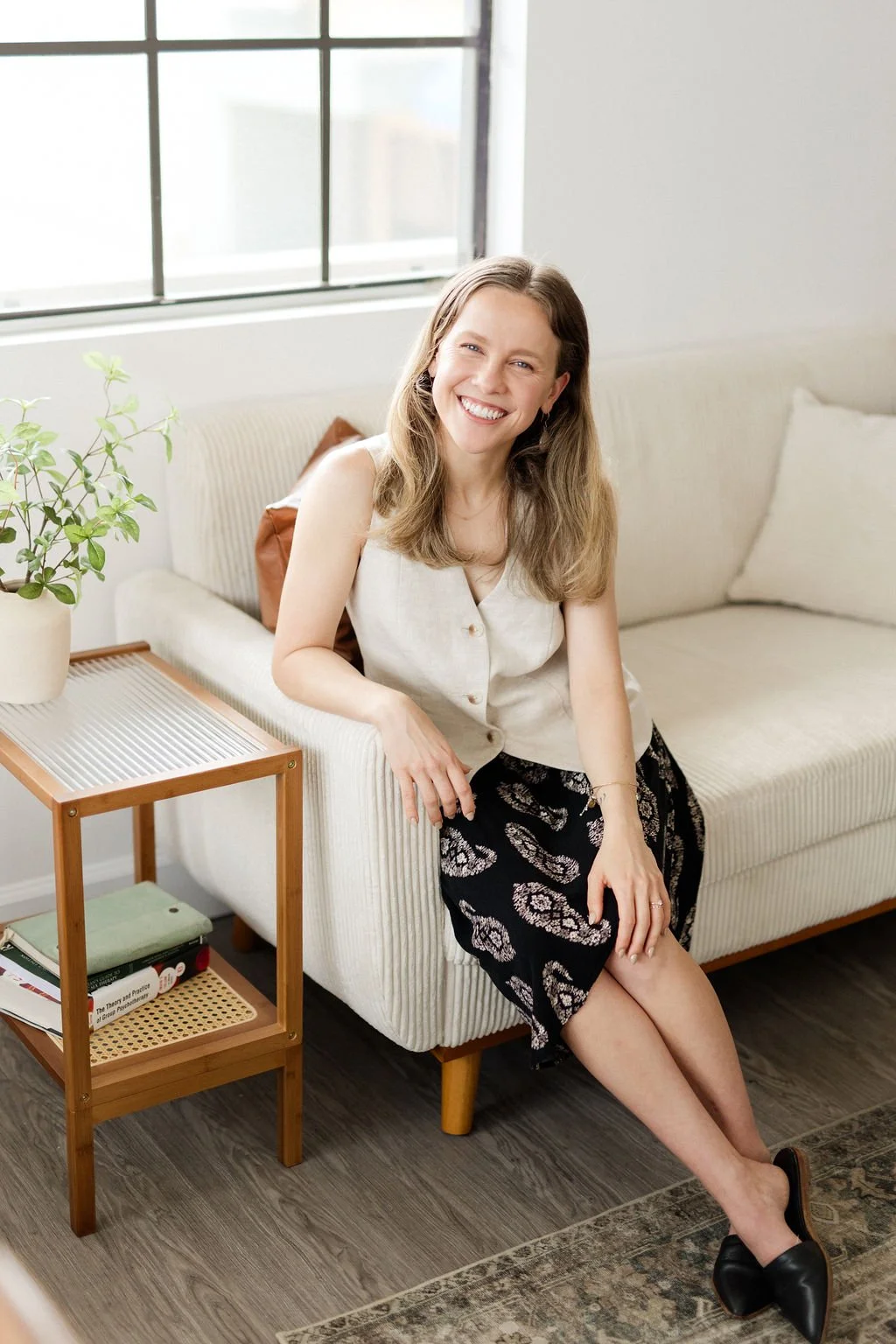 A woman with long brown hair, smiling, sitting on a cream-colored sofa in a bright room with a large window, a potted plant on a side table, and books underneath.