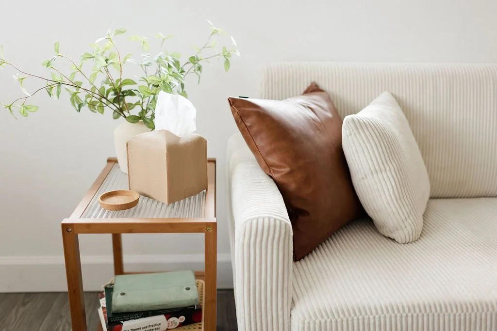 Living room with a cream striped sofa, two pillows (one white, one brown), a wooden side table with a vase holding green leafy branches, a tissue box, and a small wooden dish.