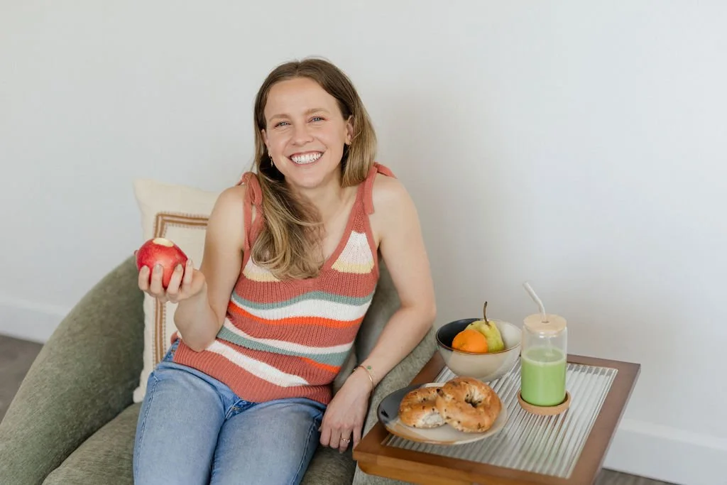 A woman with long wavy hair, smiling, sitting on a green couch, holding a red apple, wearing a colorful striped tank top and blue jeans. Next to her on a small wooden table are some bagels, a bowl of fruit, and a green beverage.