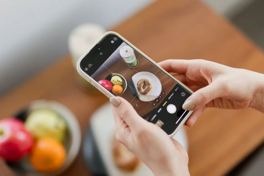 Person taking a photo of apples and a donut with a smartphone on a wooden table.