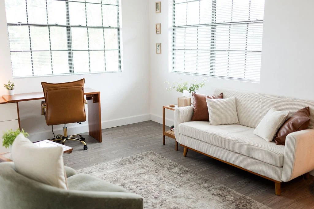 Bright living room with white walls, large windows with blinds, a white sofa with multiple throw pillows, a small side table with plants and decorative items, a wooden desk with a brown chair, and a light-colored armchair.