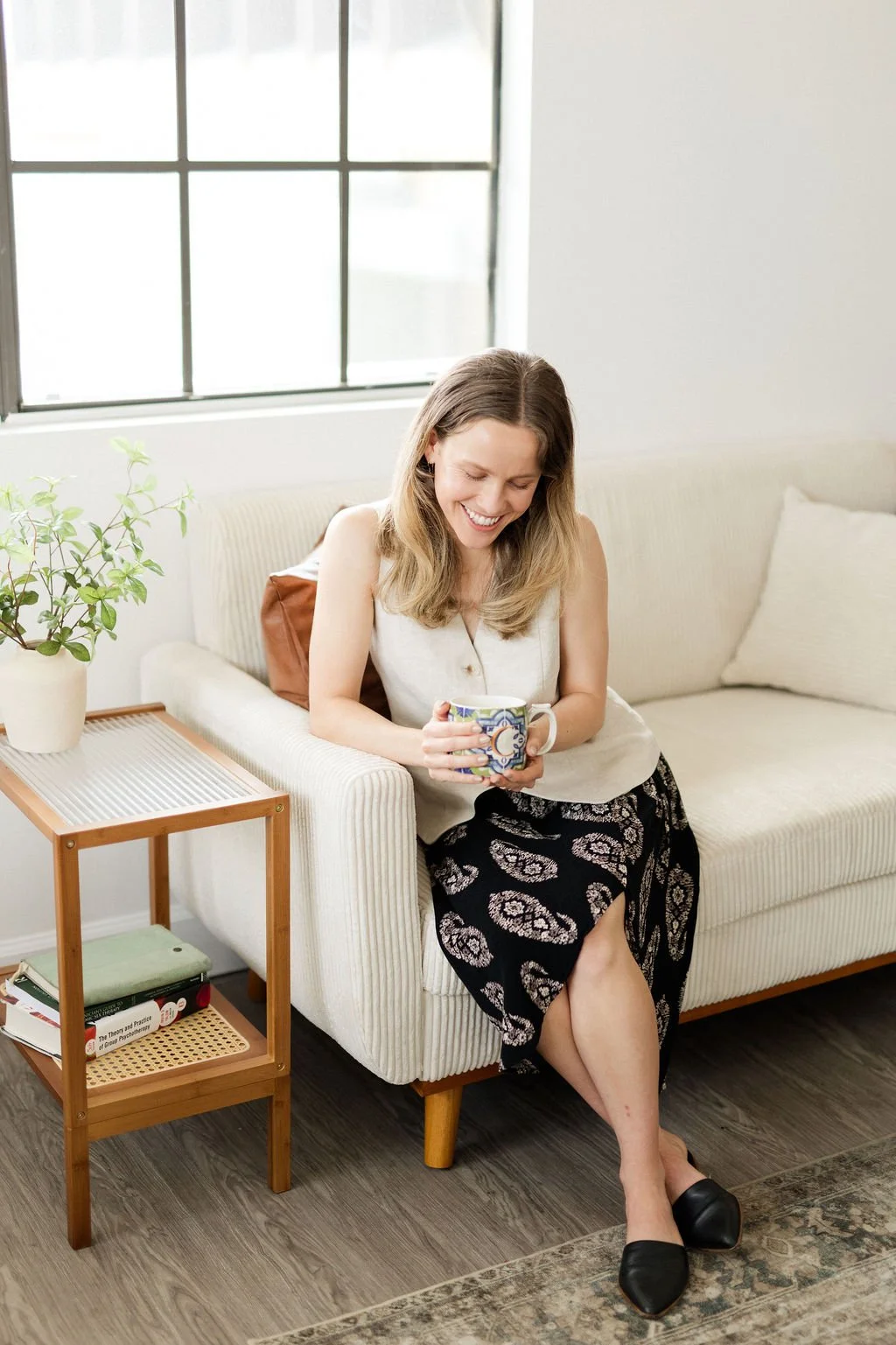 A woman with long brown hair sitting on a white sofa, smiling and holding a colorful mug. She is wearing a white sleeveless top, a black skirt with a white floral pattern, and black flats. There is a small wooden side table with a potted plant and books beside her. A large window with black grid frames is behind her, allowing natural light into the room.