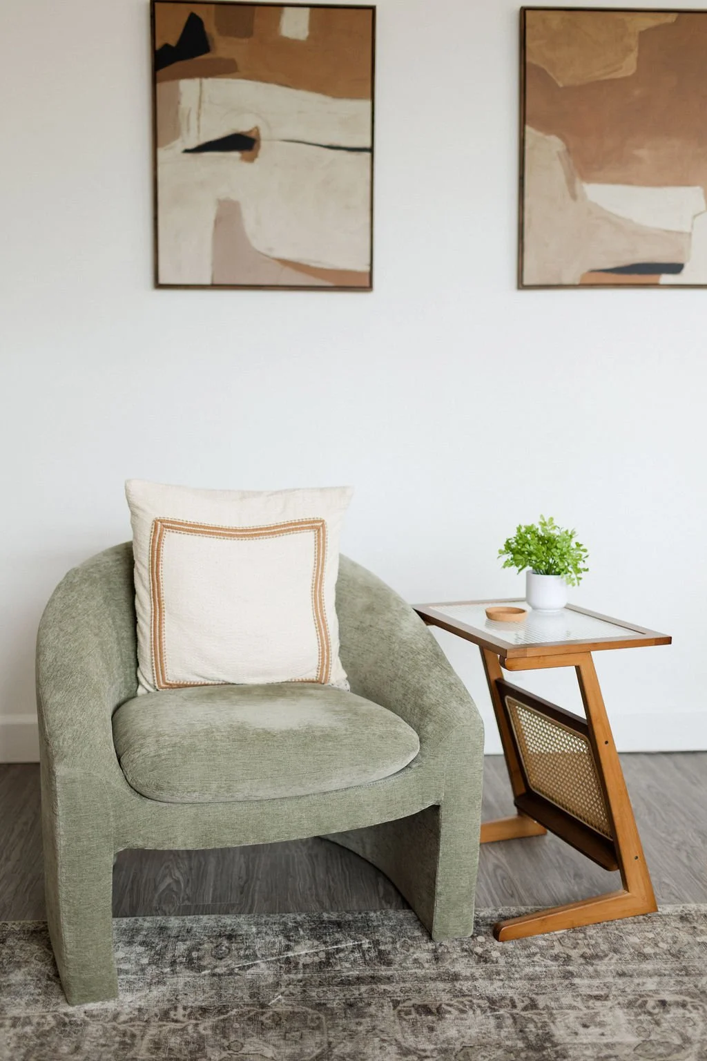 A cozy living room corner with an olive green upholstered armchair featuring a cream and beige throw pillow, a small wooden side table with a pot of green plants, a white wall with two abstract paintings, and a patterned rug on dark wooden flooring.