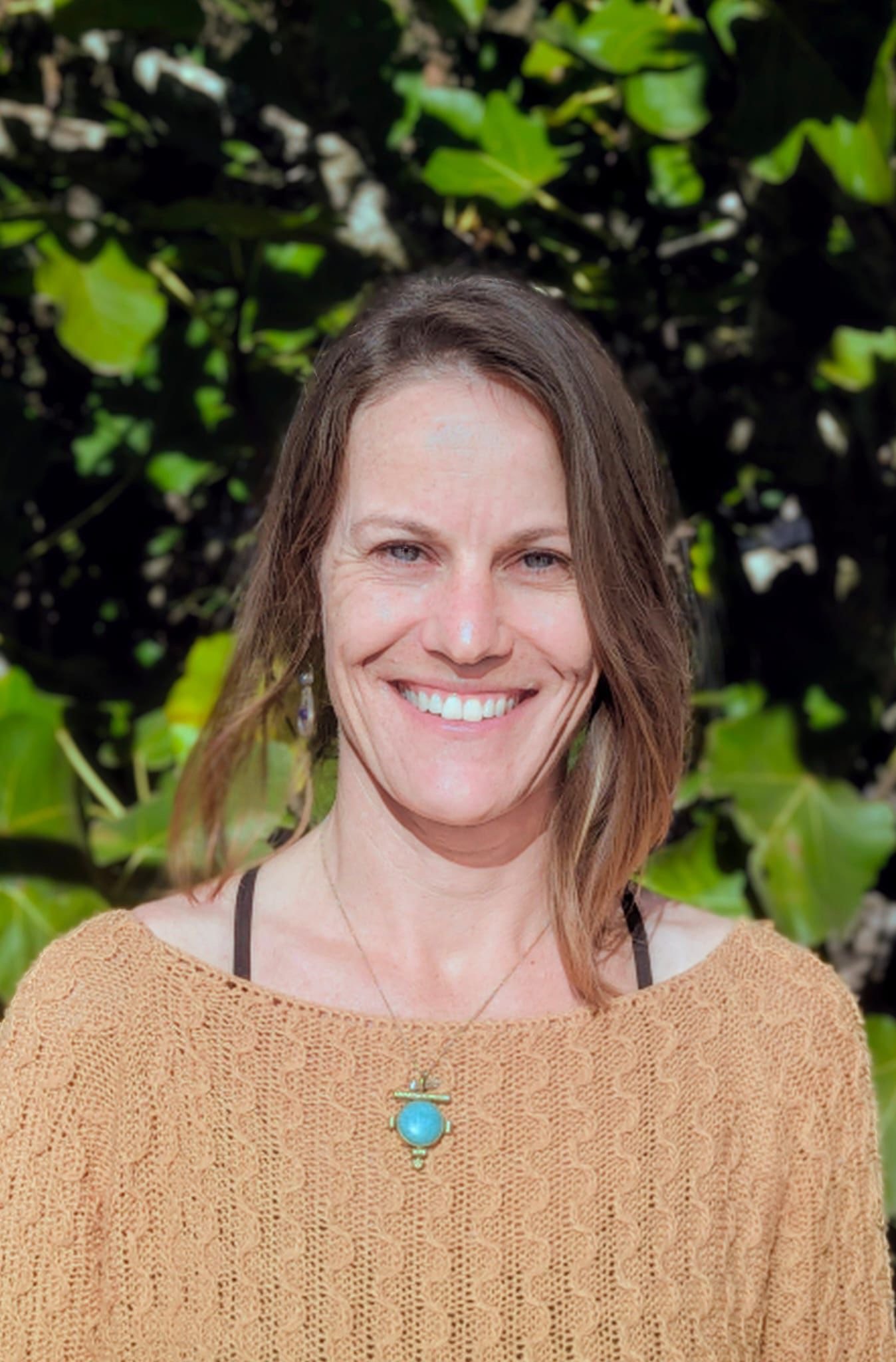 A woman with shoulder-length brown hair smiling outdoors with green leaves in the background, wearing a tan knit top and a turquoise pendant necklace.