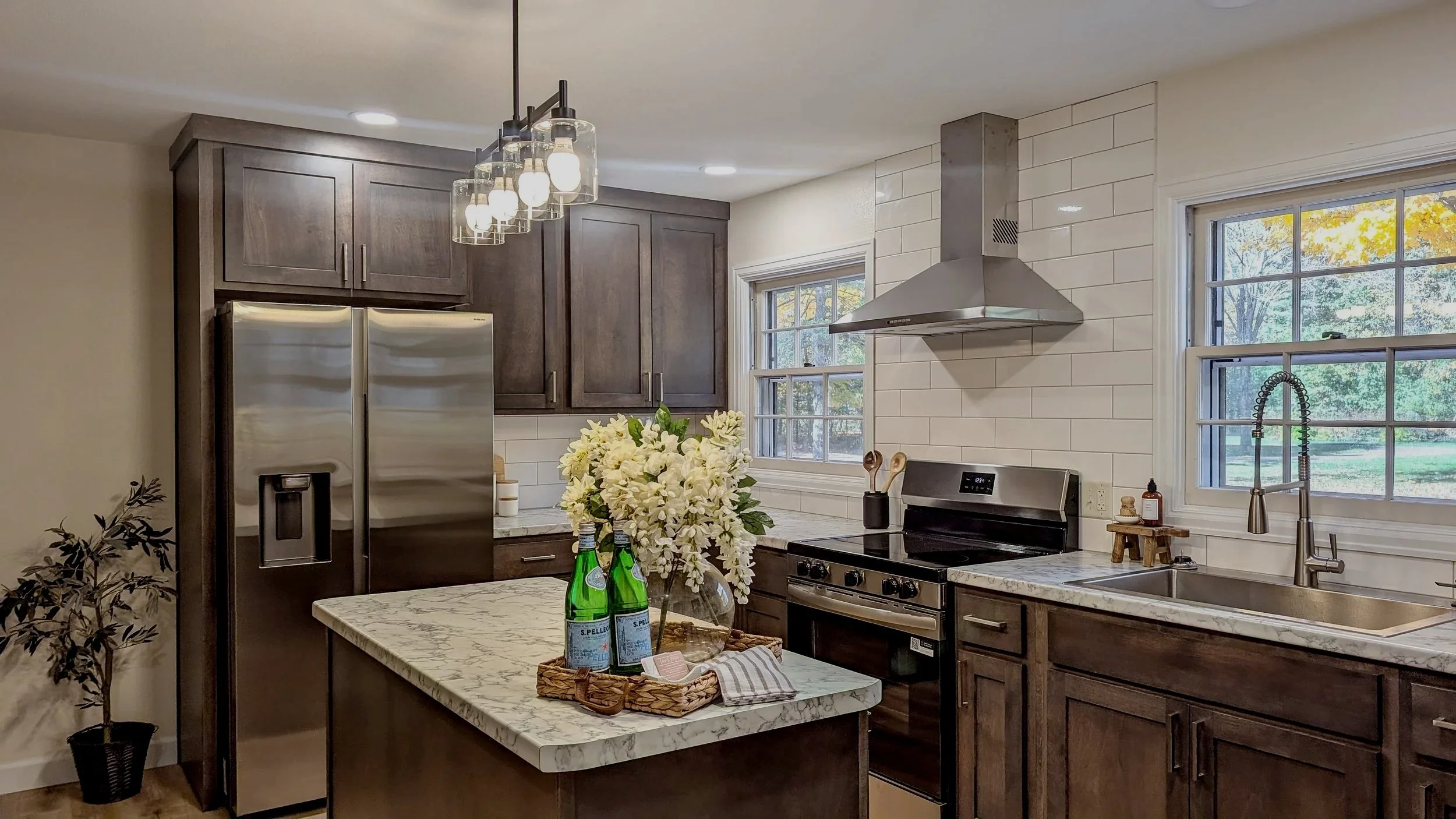 Modern kitchen with dark wooden cabinets, stainless steel refrigerator and oven, white tiled backsplash, window over the sink, marble countertop, and decorative flowers and bottled water on the island.