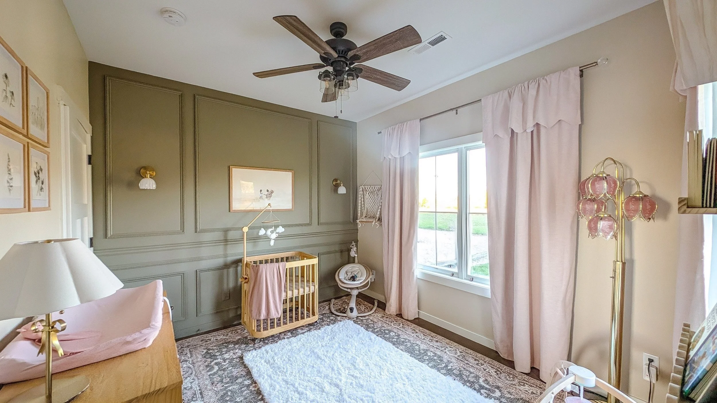 A nursery room with green and cream walls, a ceiling fan, pink curtains, a white fluffy rug, and various baby furniture and decorations.