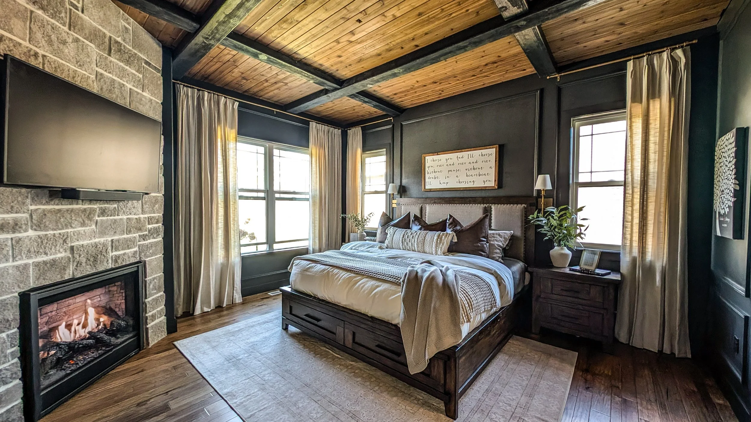 A cozy bedroom with a wood-paneled ceiling, a stone fireplace with a mounted TV, and large windows with cream curtains. The bed has dark wood frame, beige bedding, and multiple pillows. There are nightstands with lamps and plants on either side of the bed, and a framed quote hangs above the headboard.
