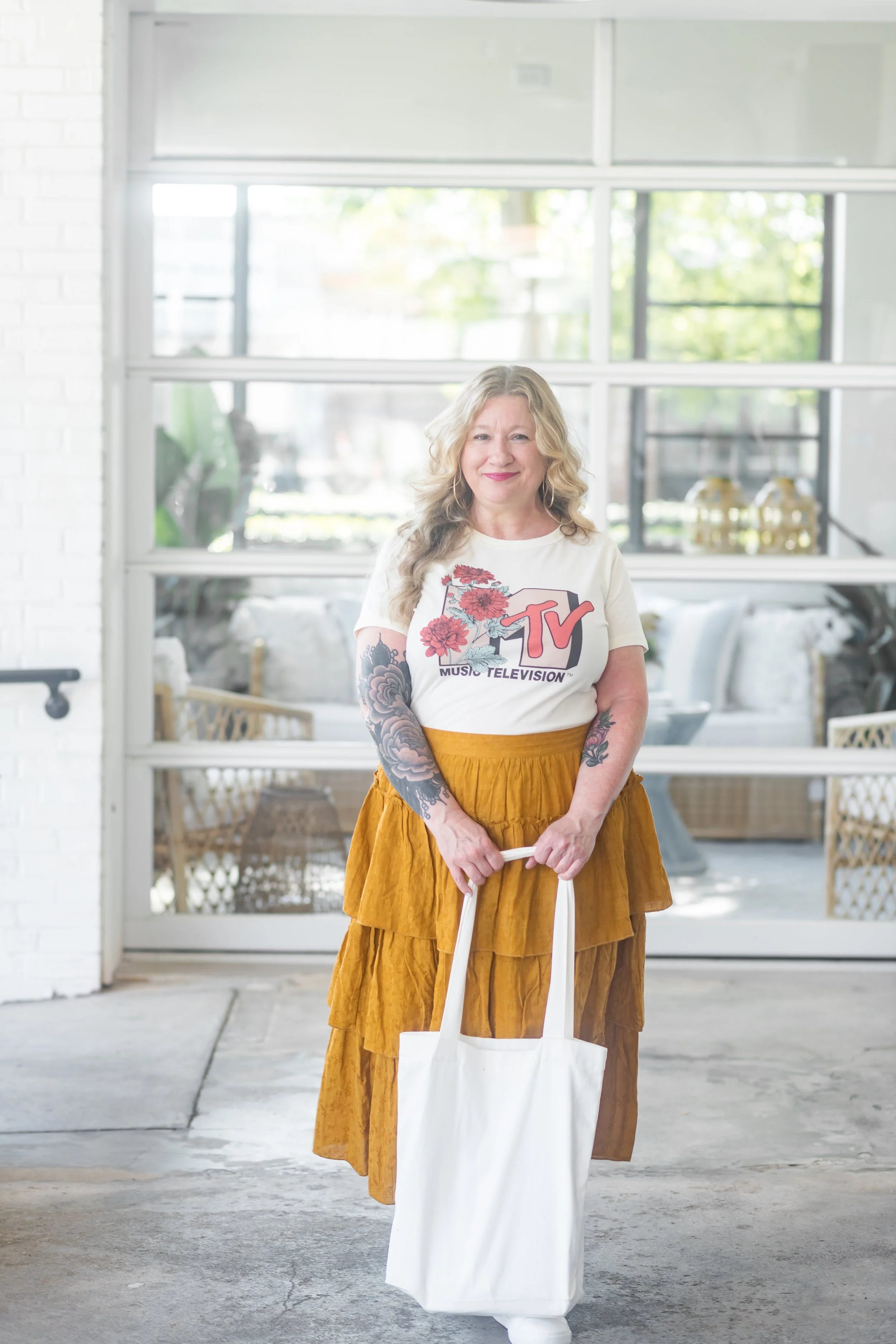 A woman with long blonde curly hair, wearing a white T-shirt with MTV logo and flowers, a mustard yellow tiered skirt, and holding a white tote bag, standing outside in front of a glass door with white brick wall on the side.