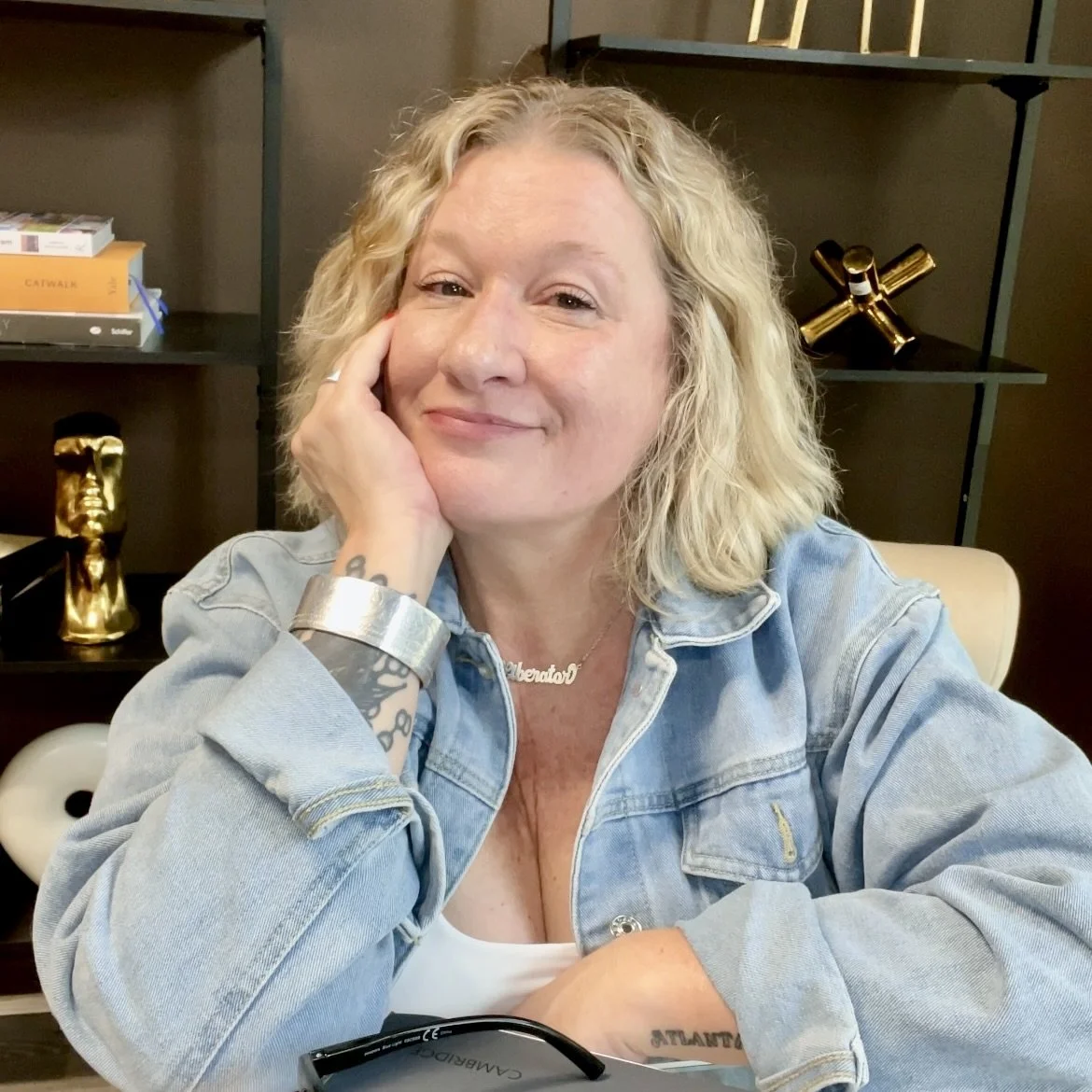 A smiling woman with blonde curly hair, wearing a denim jacket and a necklace, sitting at a table with decorative items in the background.
