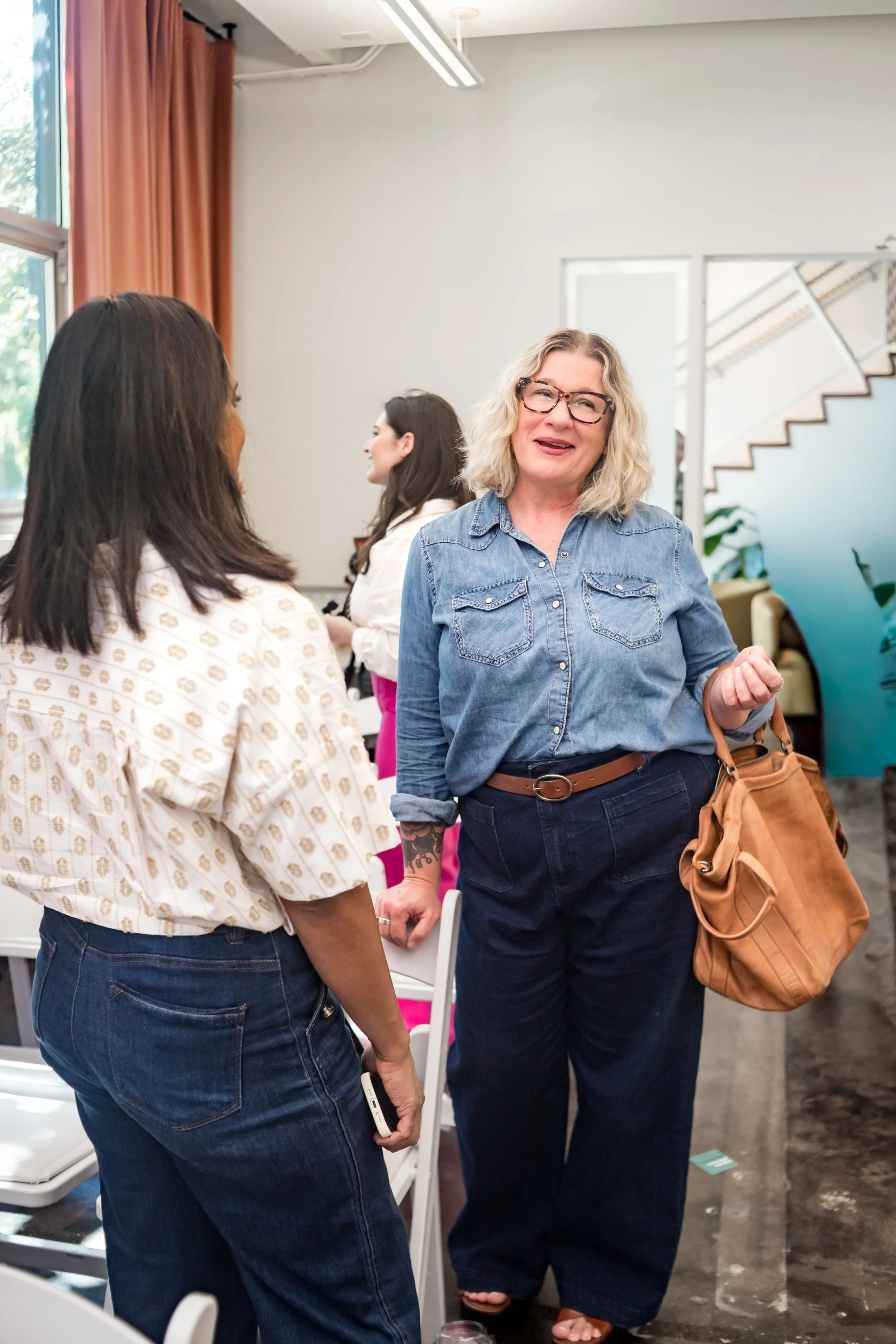 Three women are engaged in conversation indoors, with one woman holding a large tan purse and wearing glasses, denim shirt, and wide-leg jeans, while two others are partially visible.