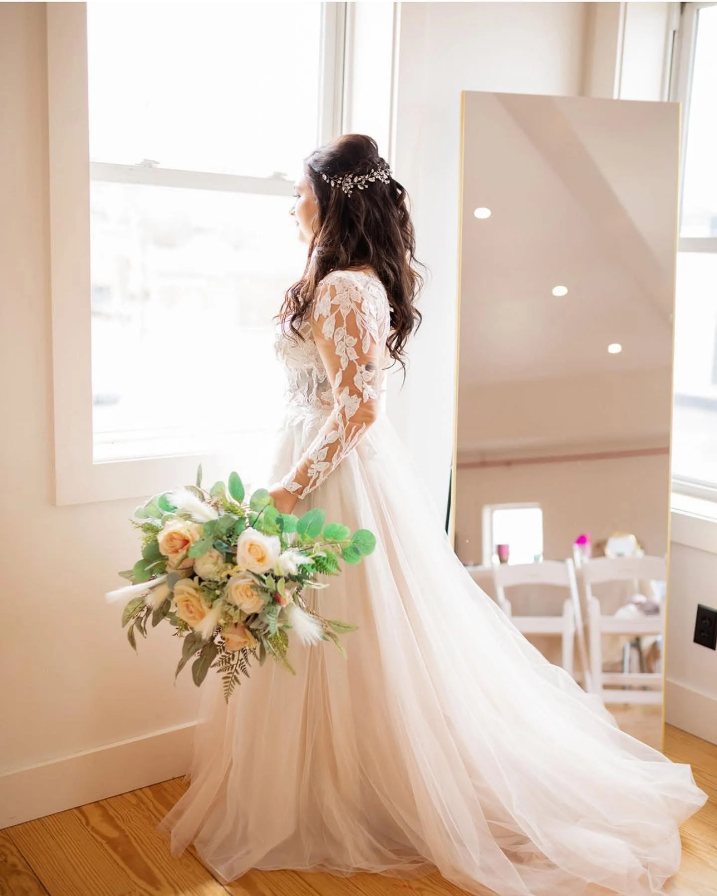 Bride in a lace wedding gown holding a bouquet of roses and greenery, facing a window, with a large mirror reflecting part of the room.