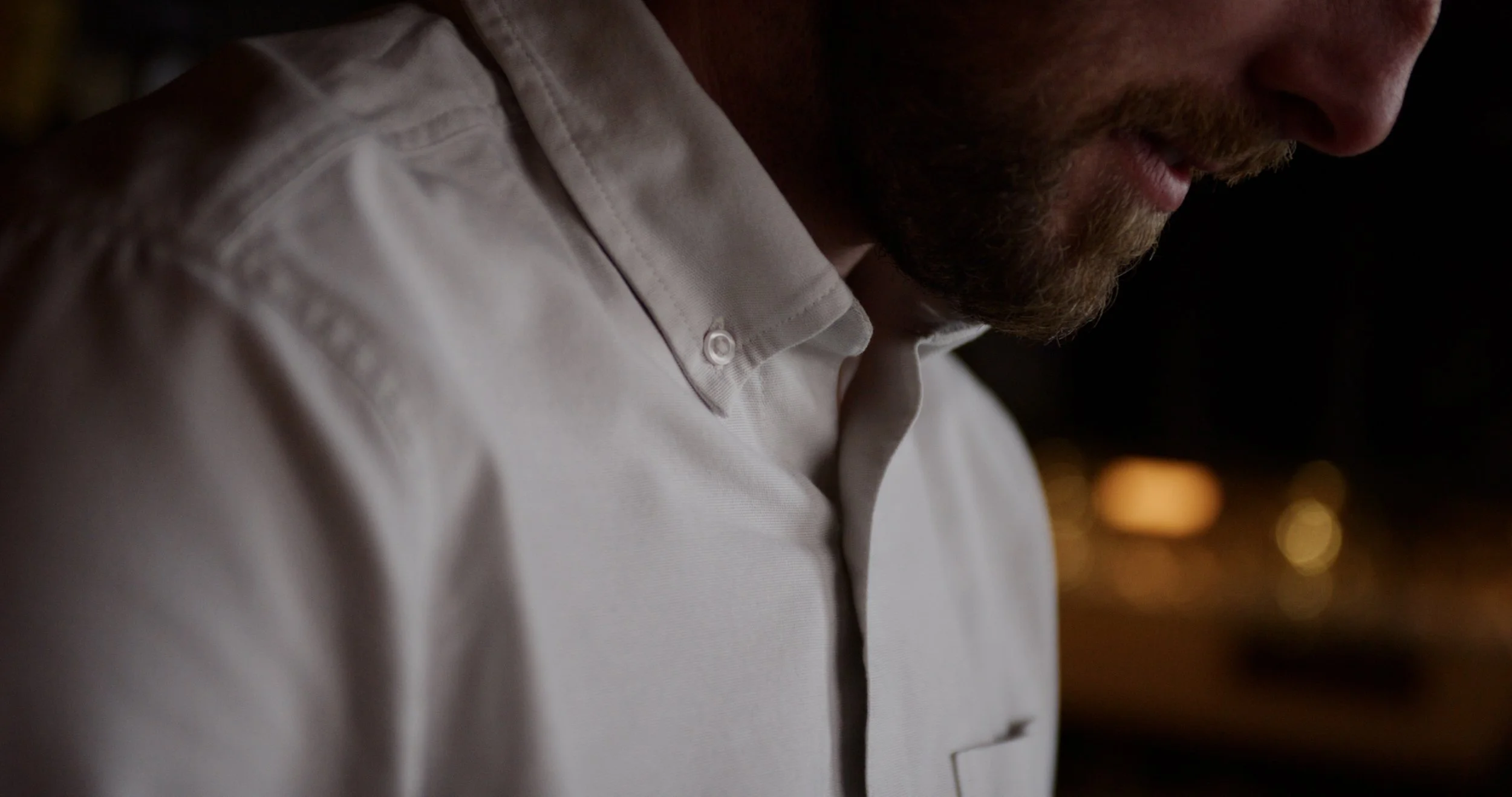 Close-up of a man wearing a light-colored dress shirt, with focus on his shoulder and neck area, at night with city lights blurred in the background.