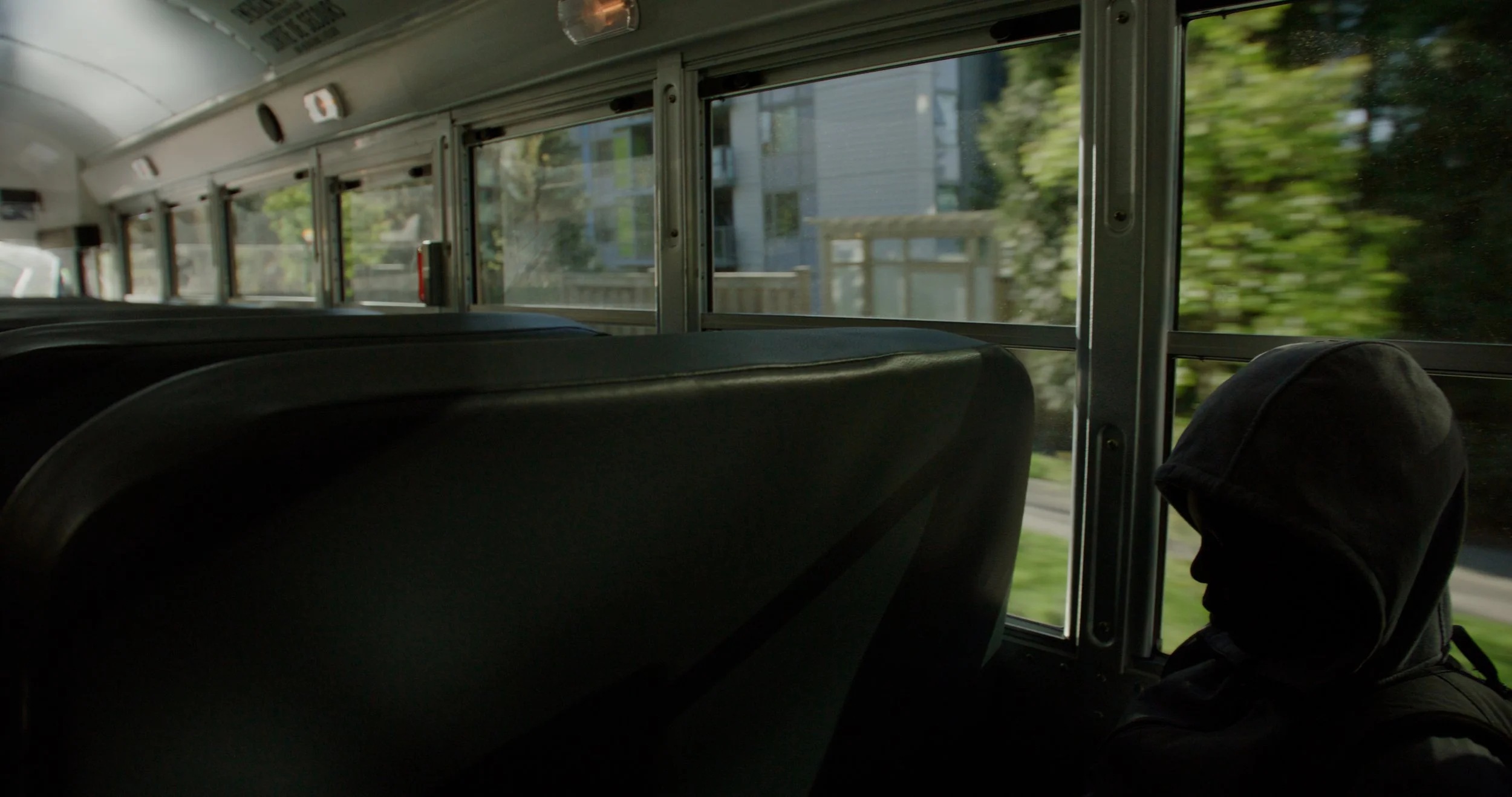 Interior of a school bus with empty black seats, a child wearing a black hooded jacket sitting quietly, and green trees and houses visible through the windows.