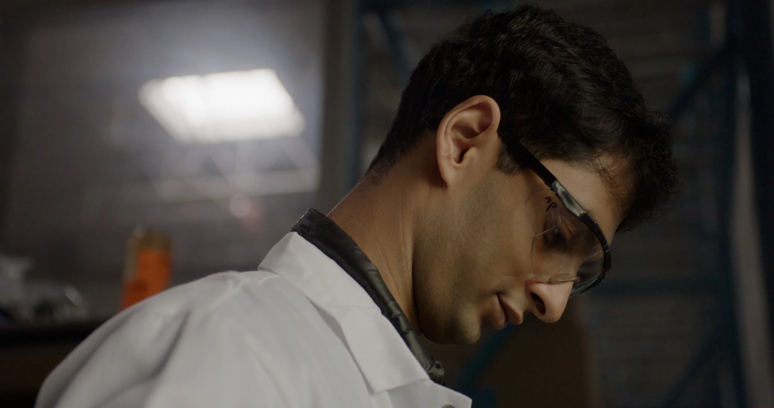 A young man wearing glasses and a white lab coat, looking down in a laboratory or industrial setting.